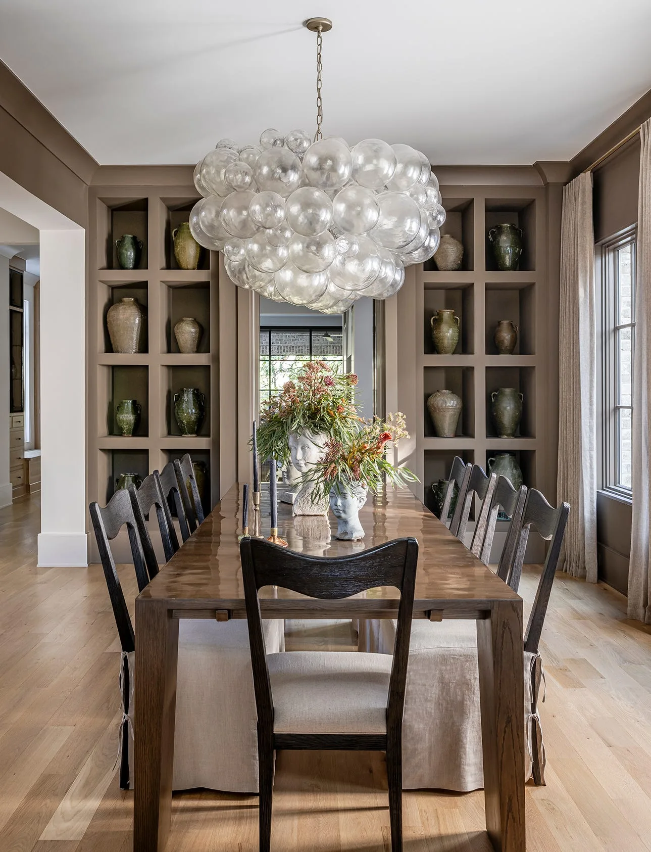 A dining room with a long wooden table, dark chairs, a large glass bubble chandelier, and symmetrical taupe built-in shelves displaying pottery.