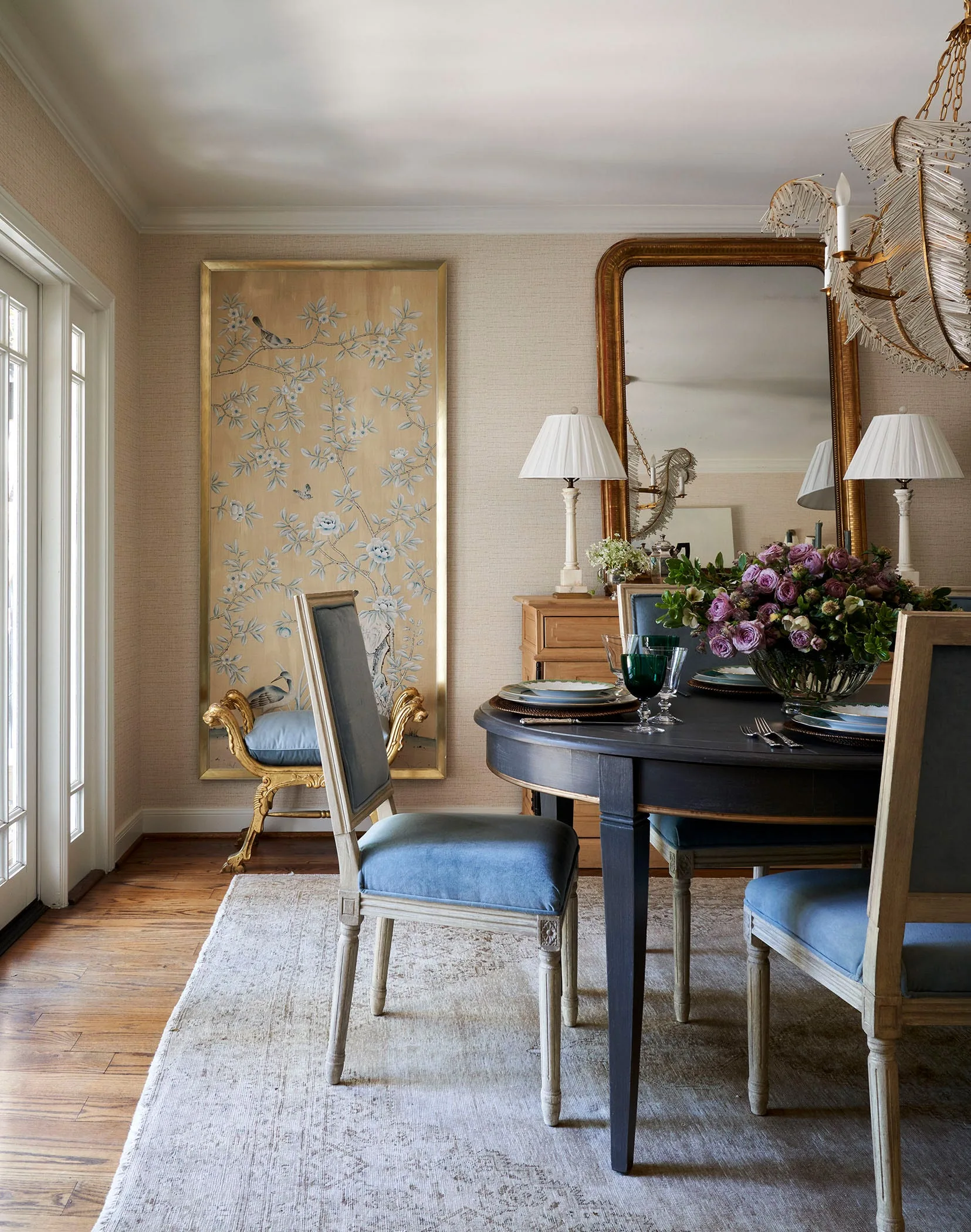 A formal dining room features blue upholstered chairs around a dark wood table with a large framed screen and mirror in the background.