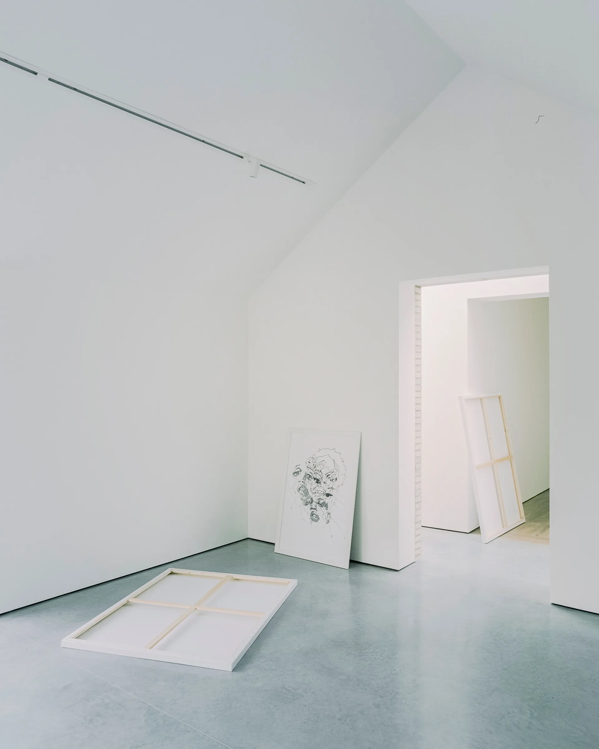 A minimalist gallery room with sloped white walls, track lighting, smooth concrete floors, and several art canvases leaning against the walls.