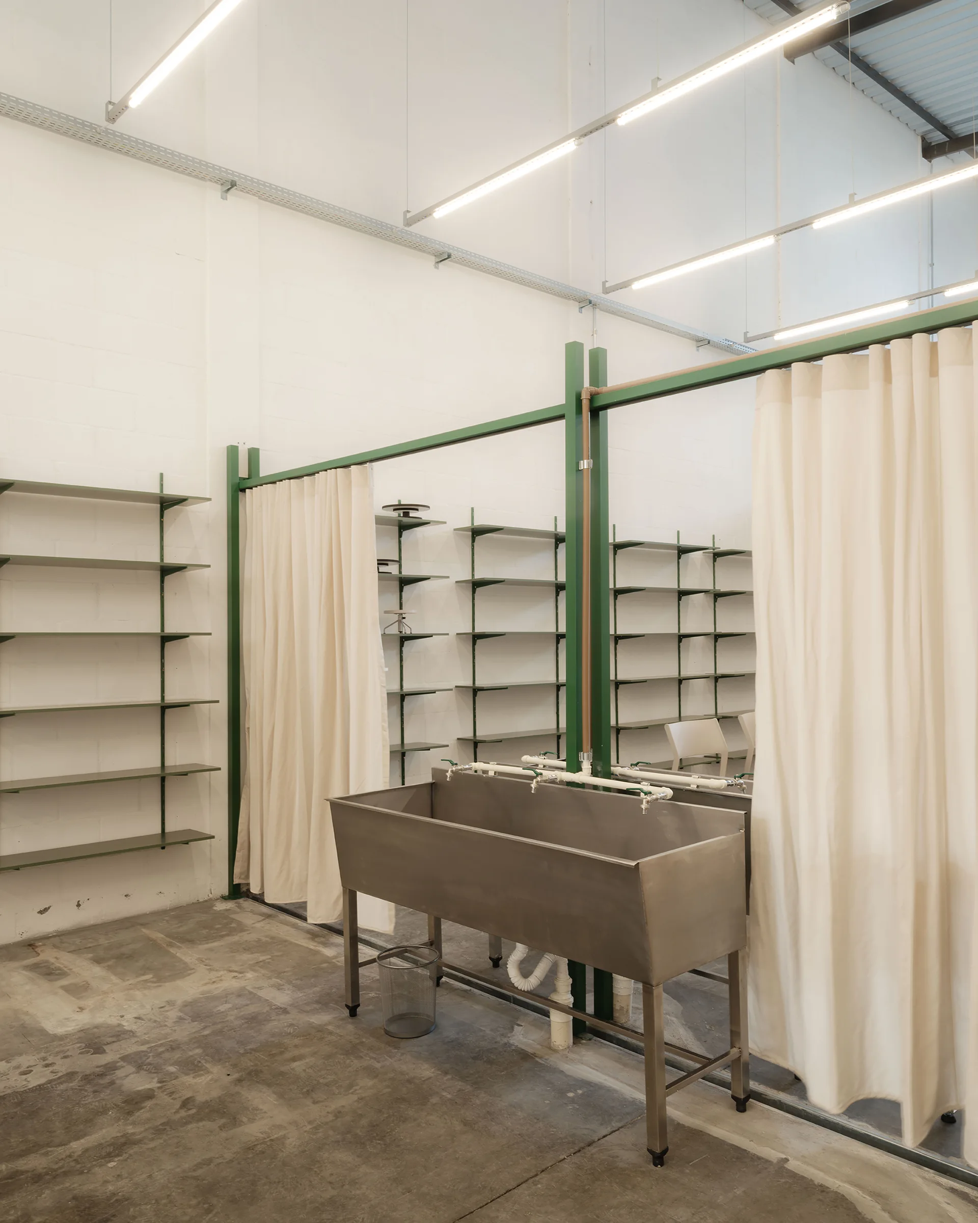 An industrial utility room features a large stainless steel sink beneath green metal posts holding cream curtains, flanked by open wall shelves.