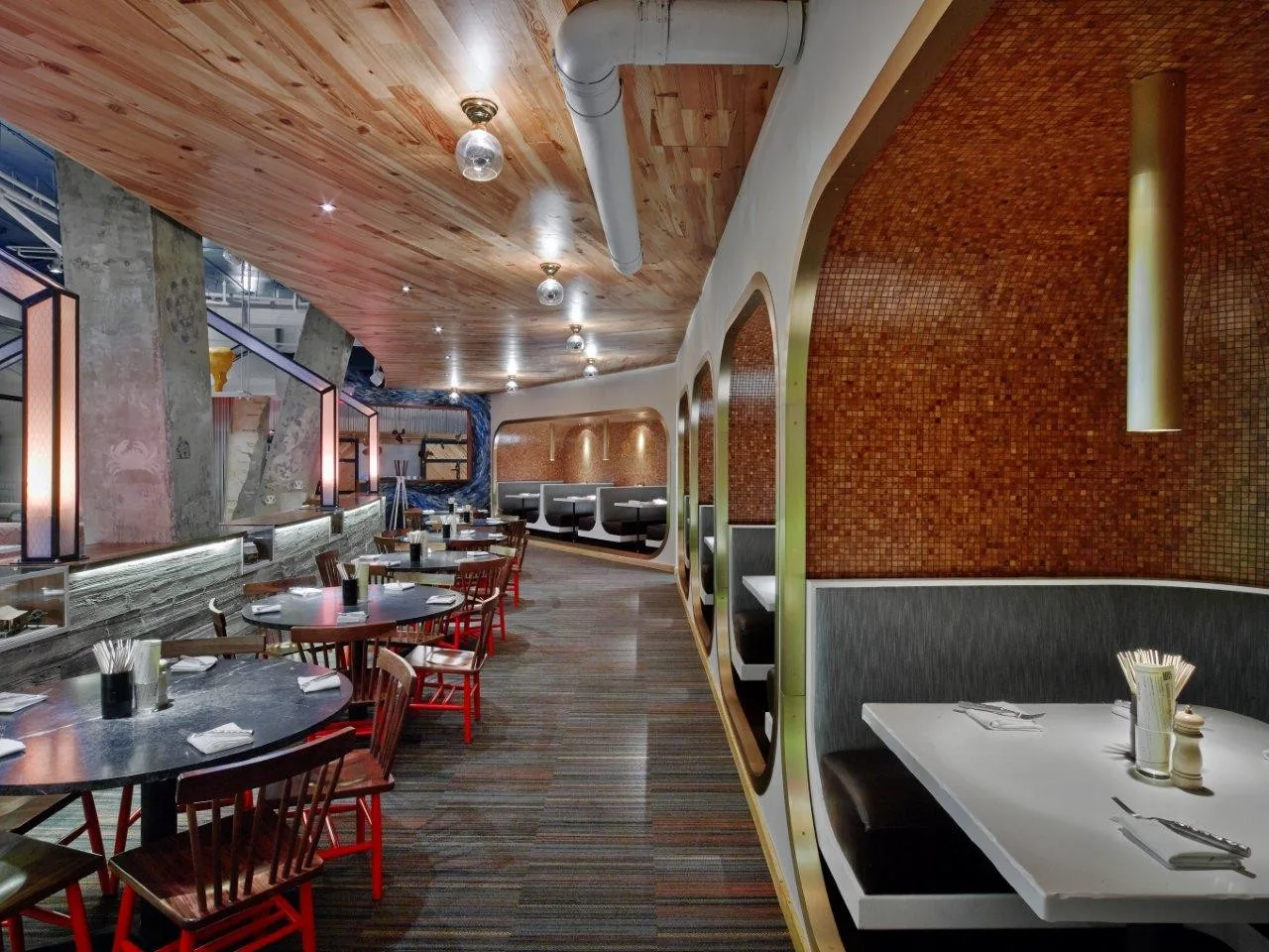 A modern restaurant dining room featuring curved wooden booths lined with mosaic tiles, round tables, and a natural wood plank ceiling.