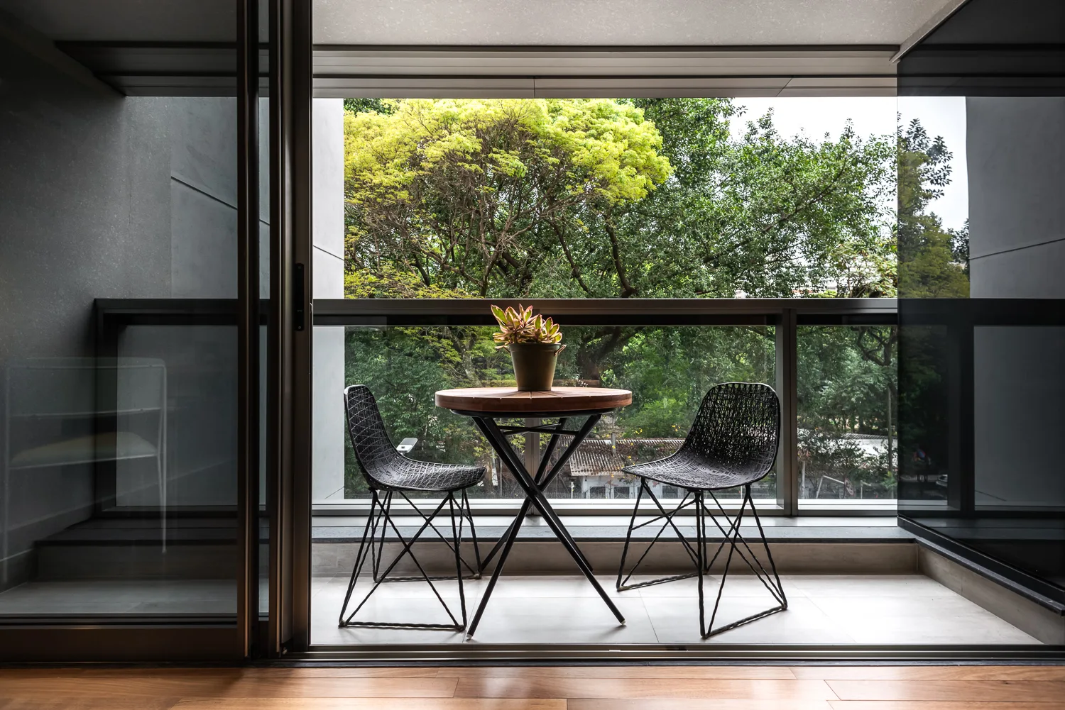 A minimalist apartment balcony featuring a black wire bistro set next to a glass railing overlooking green trees.