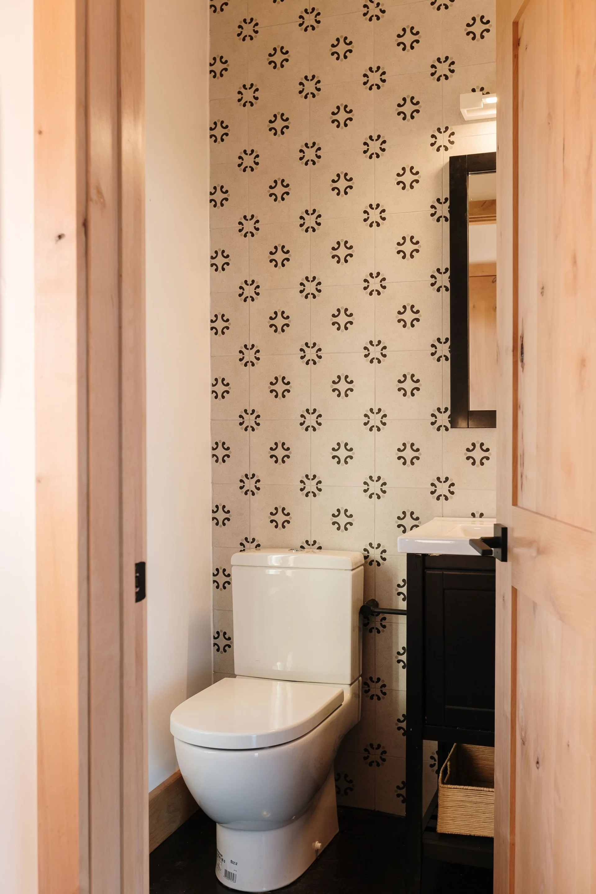 A small powder room viewed through a wood doorway, featuring a white toilet, black vanity, and a wall of cream geometric patterned tiles.