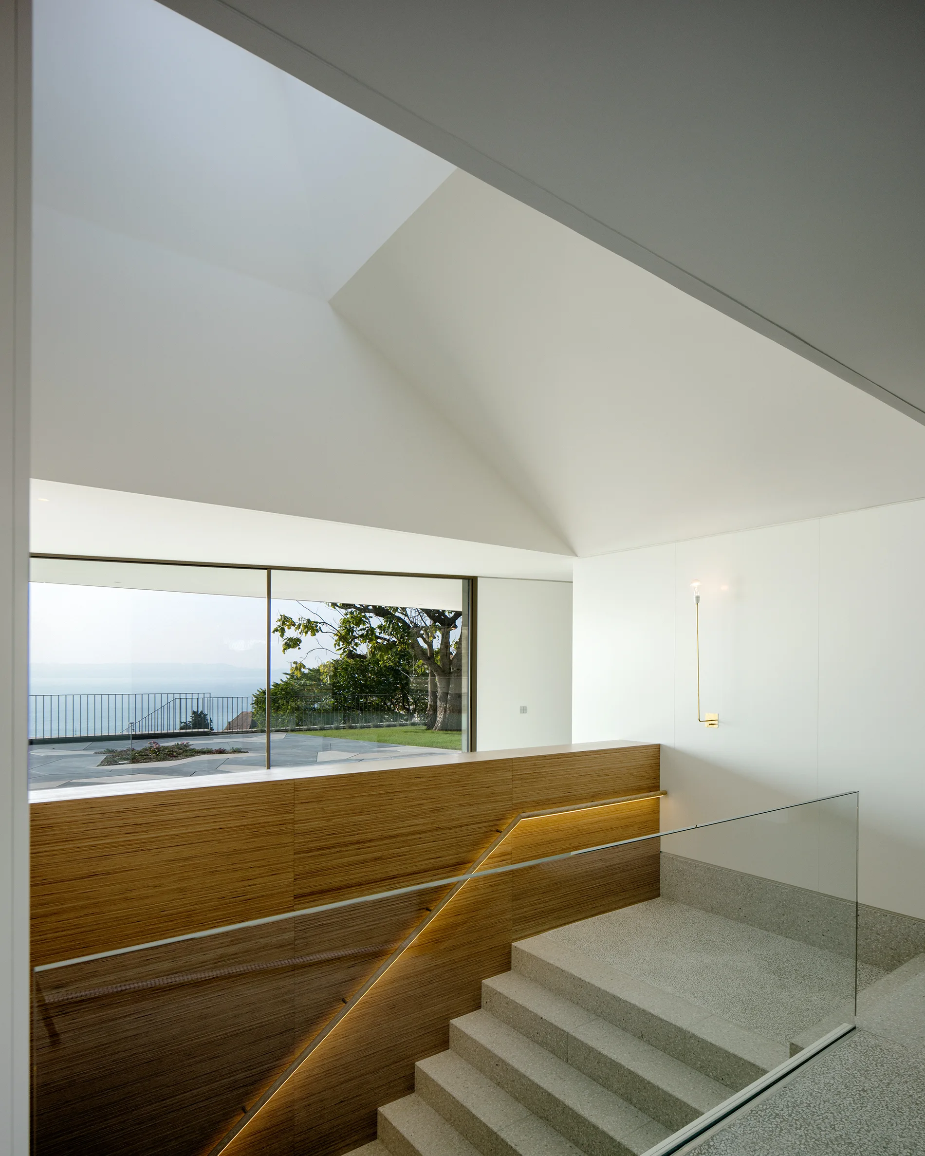 A minimalist stone staircase features a glass railing, a textured bamboo wall panel, and a large window overlooking an outdoor landscape.