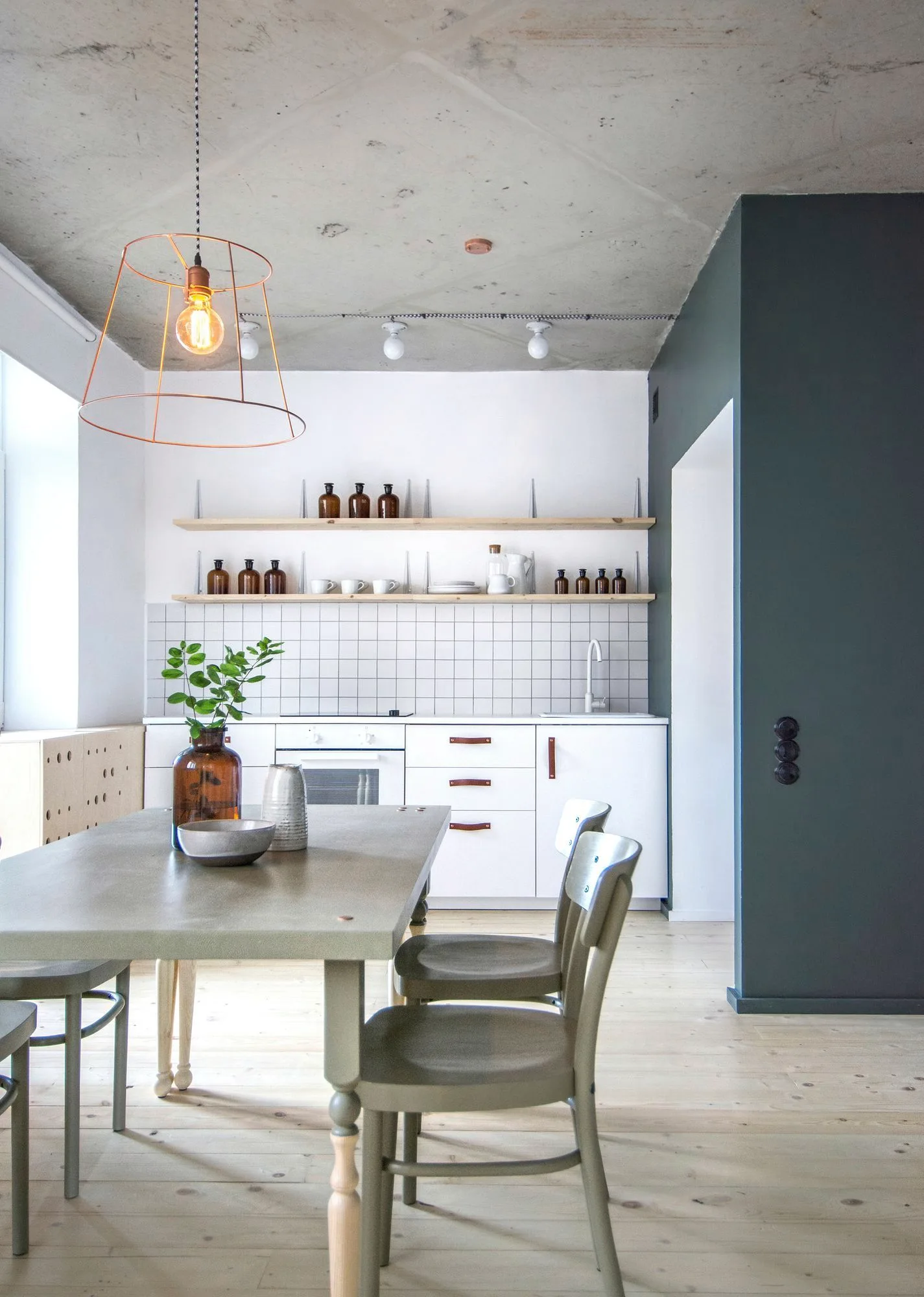 A dining table set sits before a kitchen with white cabinets, wood floating shelves, a tiled backsplash, and an exposed concrete ceiling.