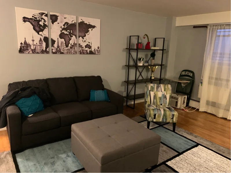 A living room features a dark grey sofa, a tufted square ottoman, a patterned accent chair, and a three-piece map canvas on a grey wall.