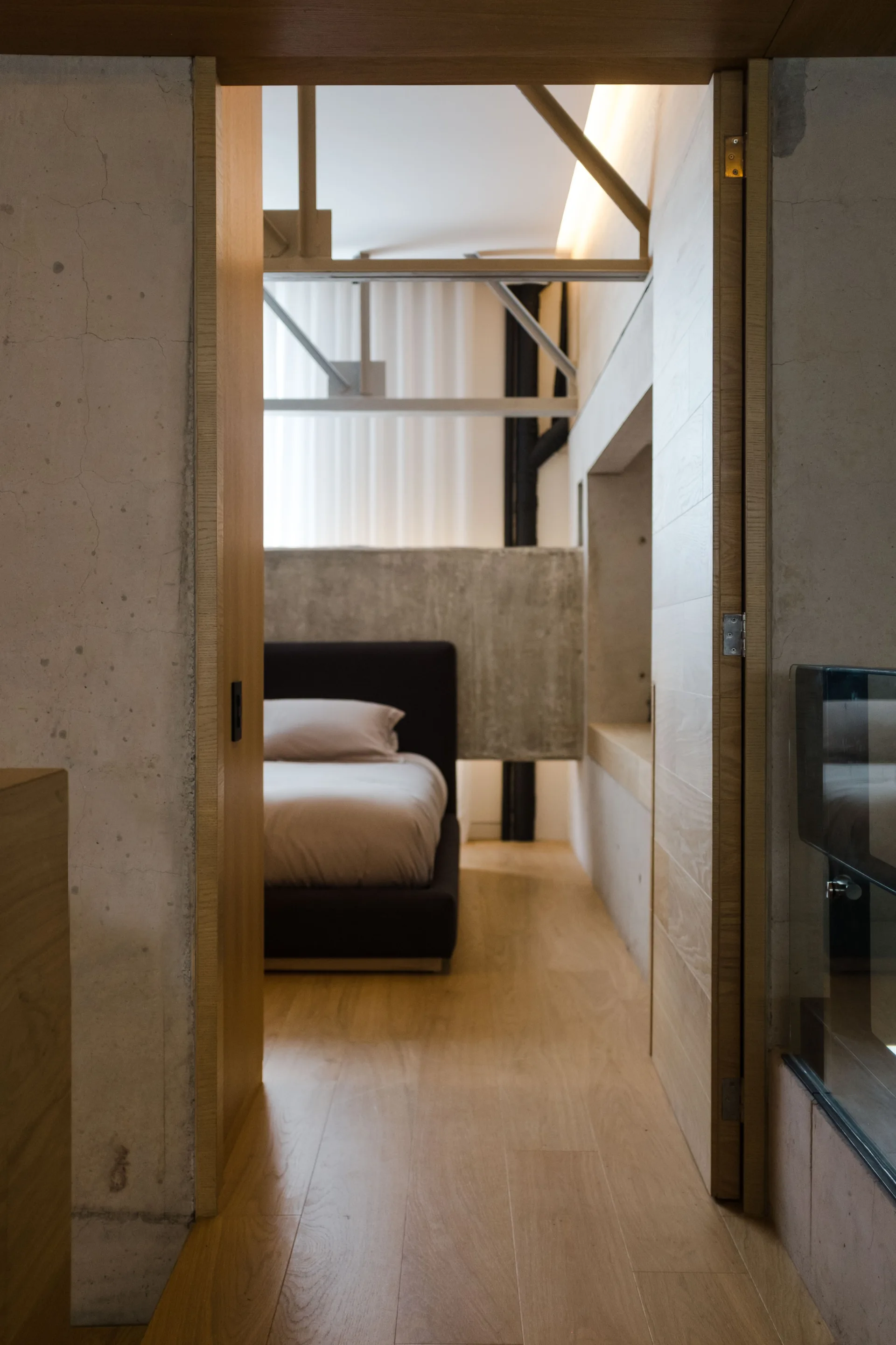 A view through a wooden doorway into a bedroom featuring a dark upholstered bed against an exposed concrete partition wall.