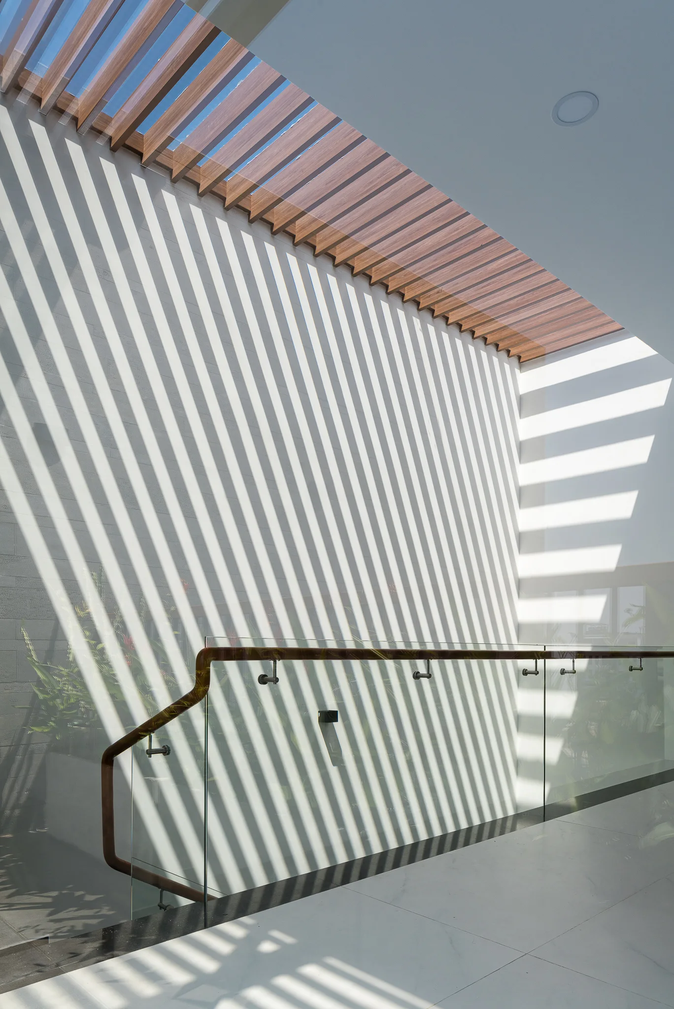 A glass staircase railing with a wood handrail stands before a white wall patterned with diagonal shadows from a slatted timber skylight.