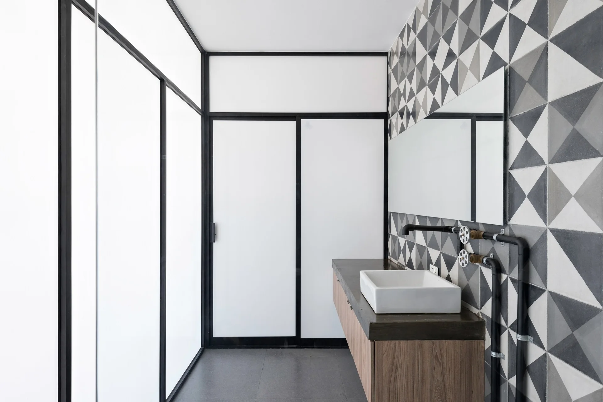 A contemporary bathroom featuring a wood-toned vanity with a white vessel sink, geometric triangle wall tiles, and black industrial pipe faucets.