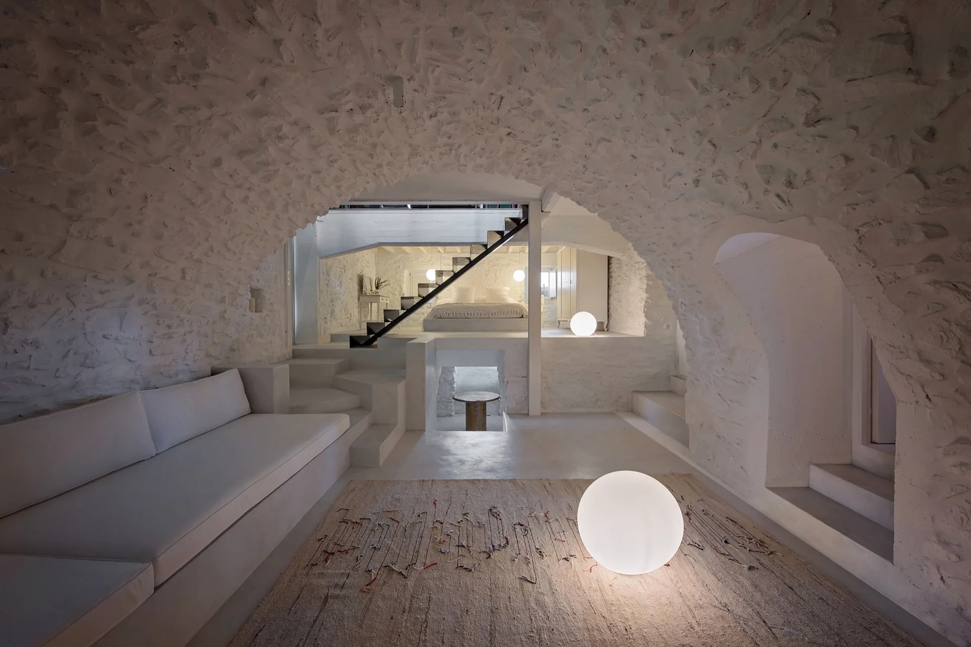 A multi-level white living area featuring textured stone vaulted ceilings, built-in plaster seating, and a staircase leading to a mezzanine bed.