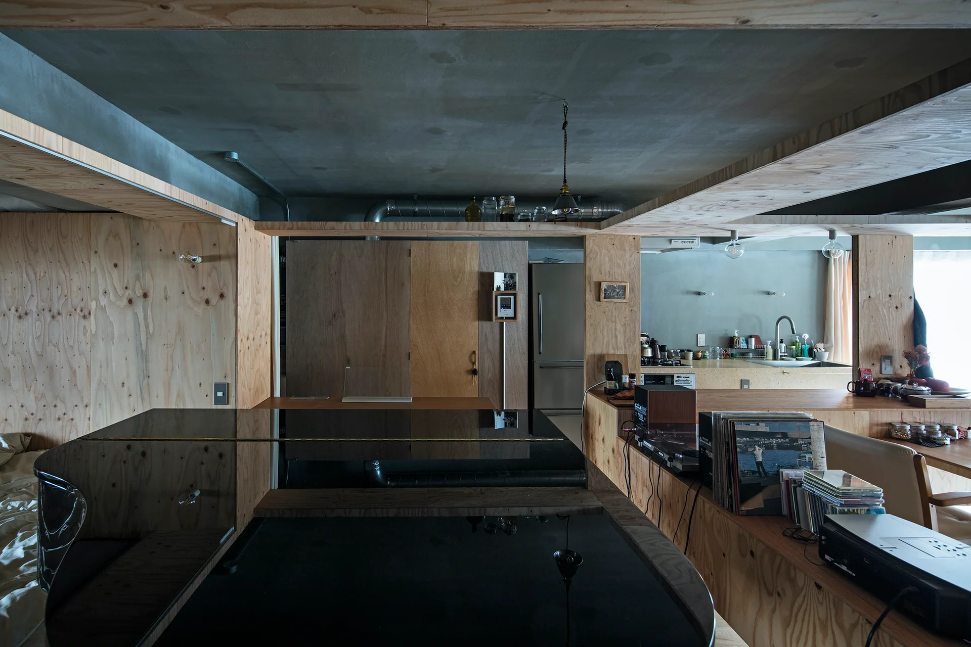 An open-concept living space features light plywood wall paneling, an exposed concrete ceiling, a black grand piano, and a kitchen in the background.