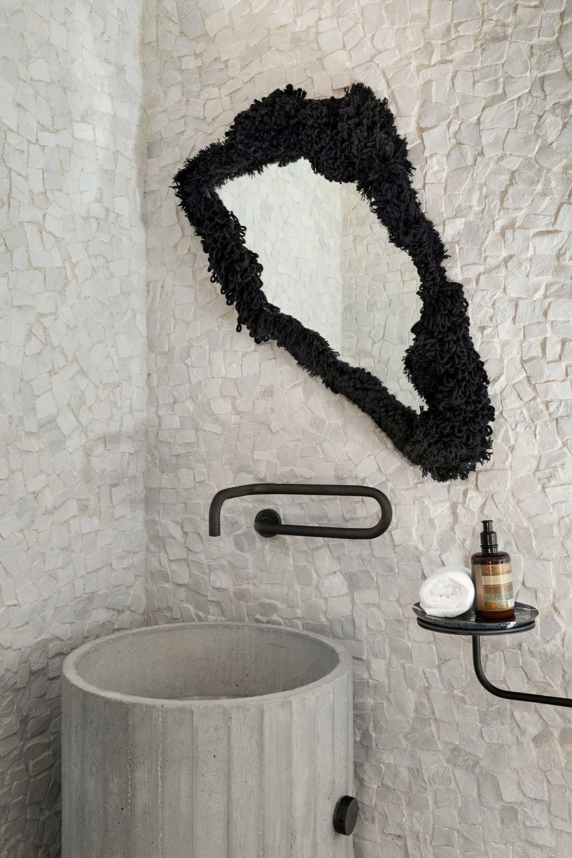 A round grey concrete pedestal sink sits below a wall-mounted faucet and an irregular mirror with a shaggy black frame on a white stone mosaic wall.