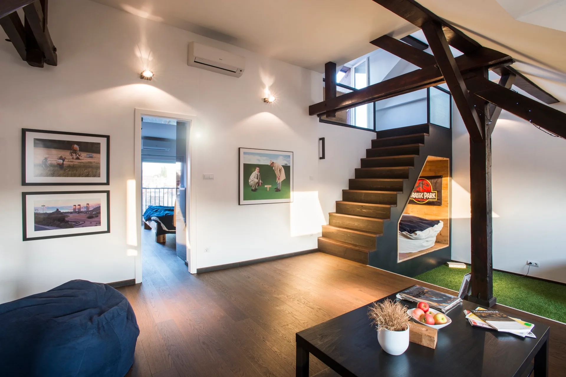 A modern loft living room features dark wood floors, exposed ceiling beams, and a dark stained staircase with an integrated sleeping nook underneath.