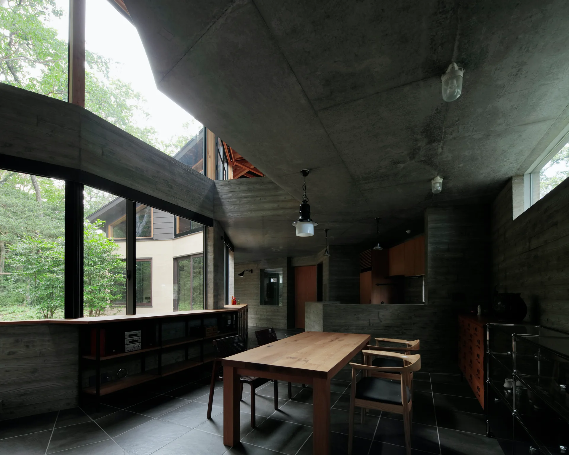 A minimalist dining room featuring a wooden dining table and chairs surrounded by textured board-formed concrete walls and large windows.