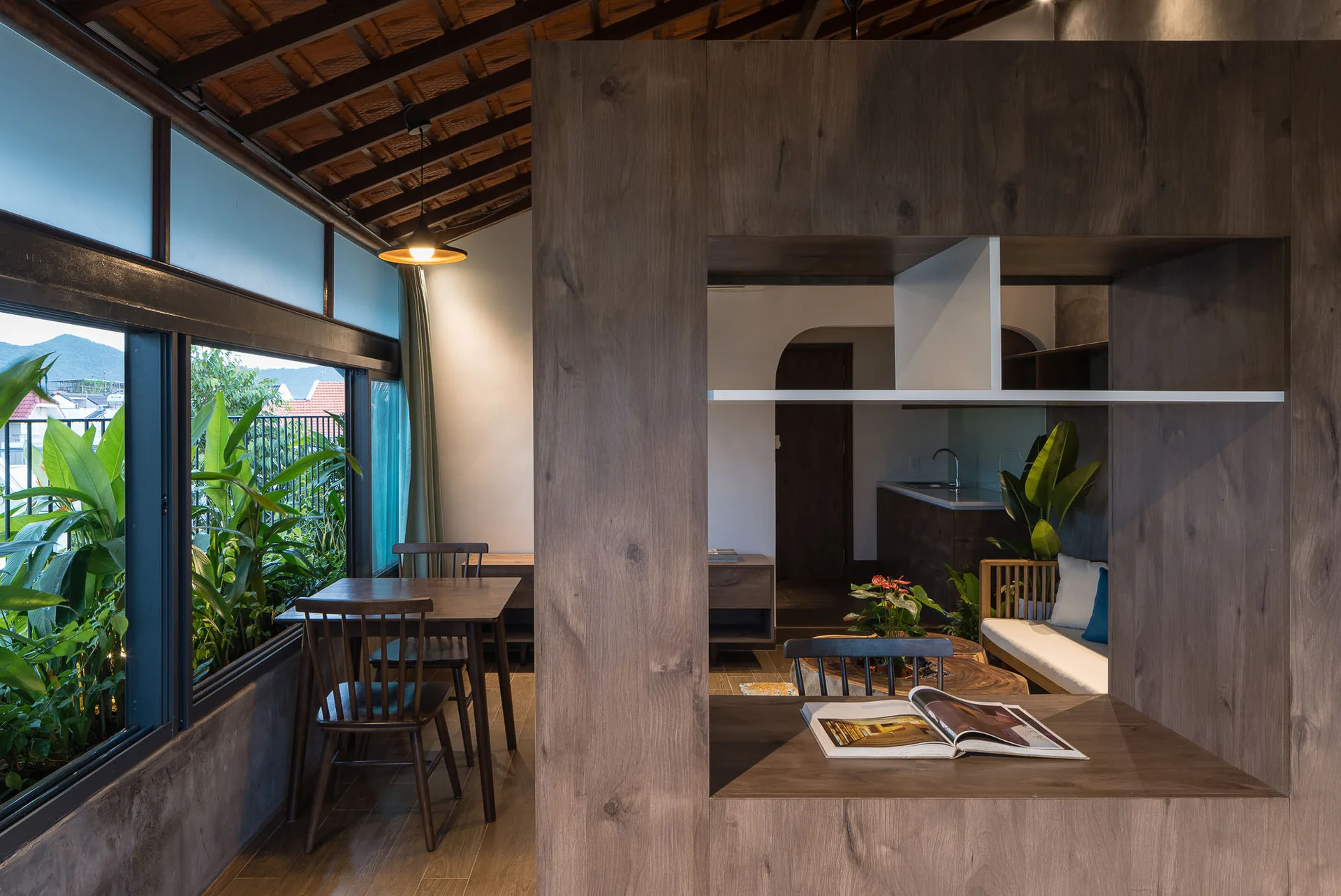 A wooden dining table sits next to a low window looking out at green plants, separated from a kitchen by a large wood room divider.