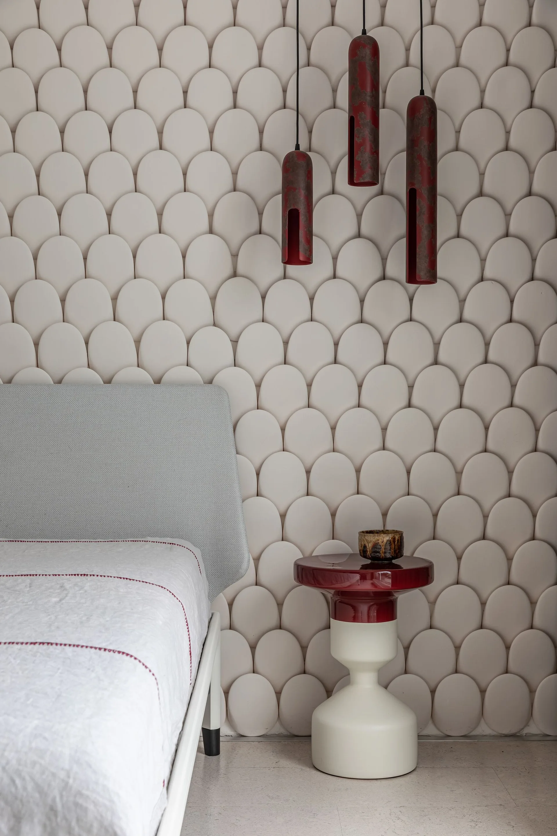 A bedroom corner shows a grey headboard beside a two-tone red pedestal side table and hanging red pendants against a white scalloped accent wall.