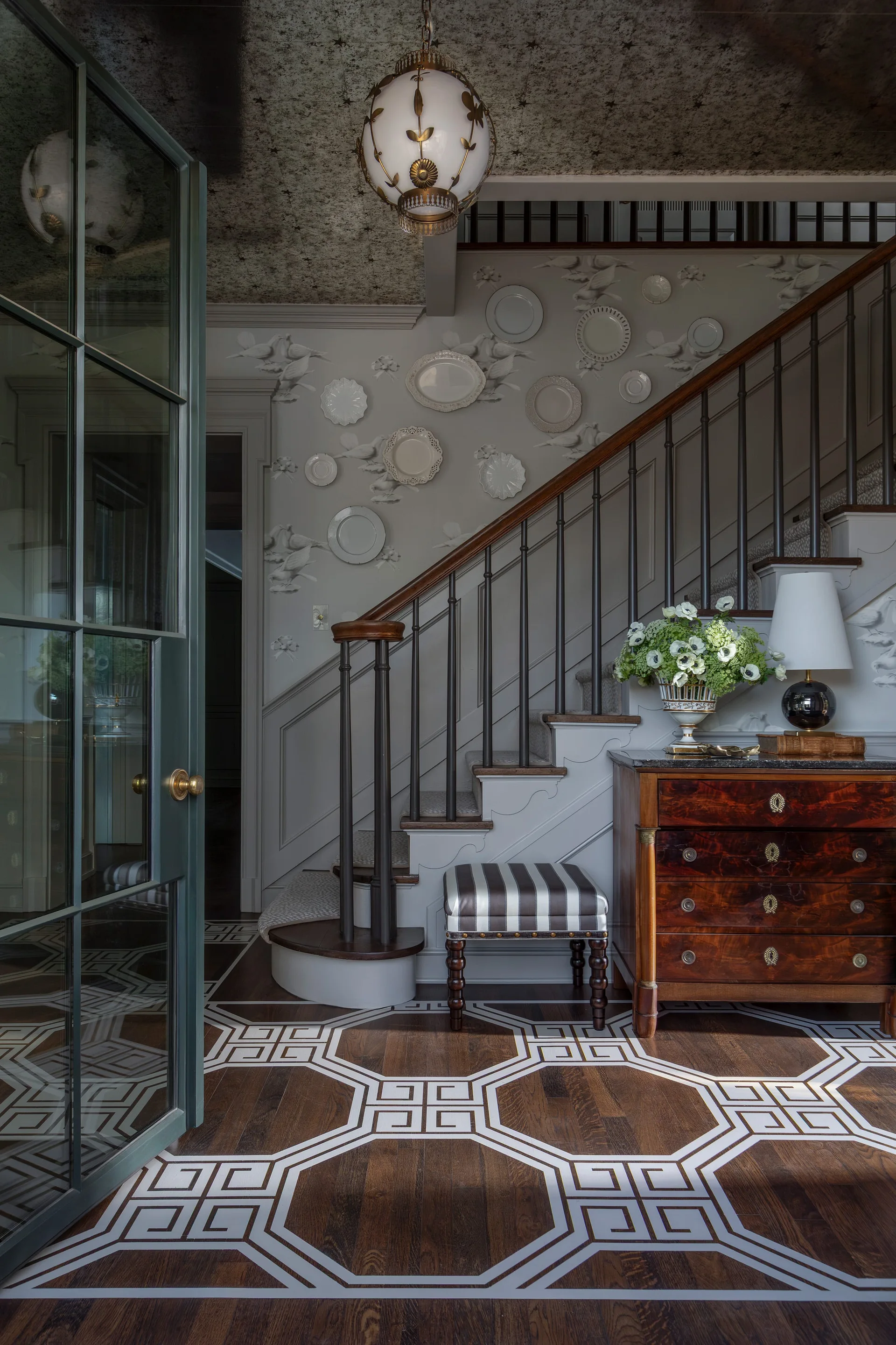 An entryway features a dark wood staircase, a wooden chest with flowers, and decorative white plates mounted on the light grey walls.