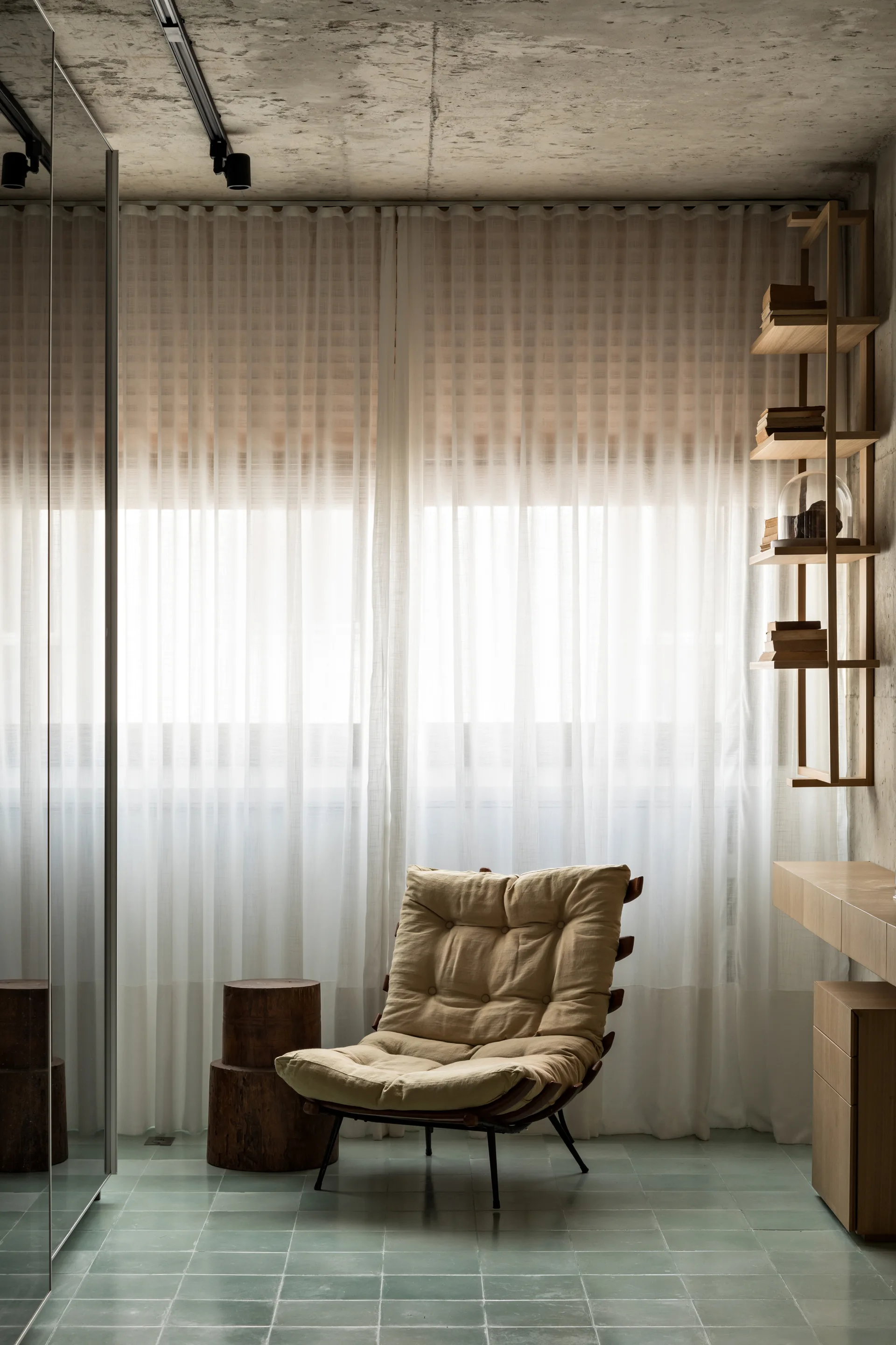 A tufted beige armchair and a log side table sit on green tiled floors in a room with a concrete ceiling and sheer curtains.