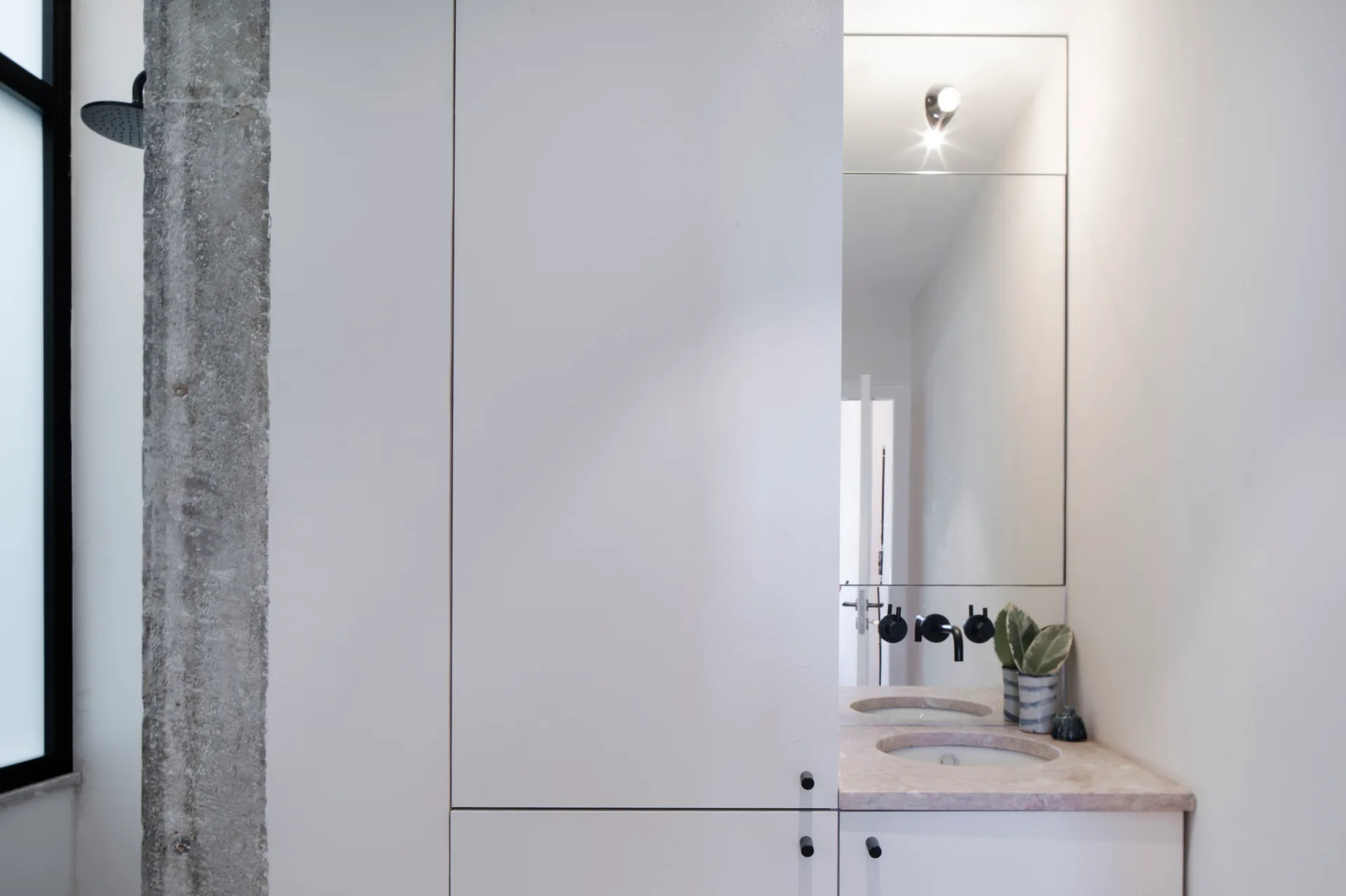 A minimalist bathroom features white cabinetry, a pinkish stone vanity with integrated sinks, black faucets, and an exposed concrete pillar.