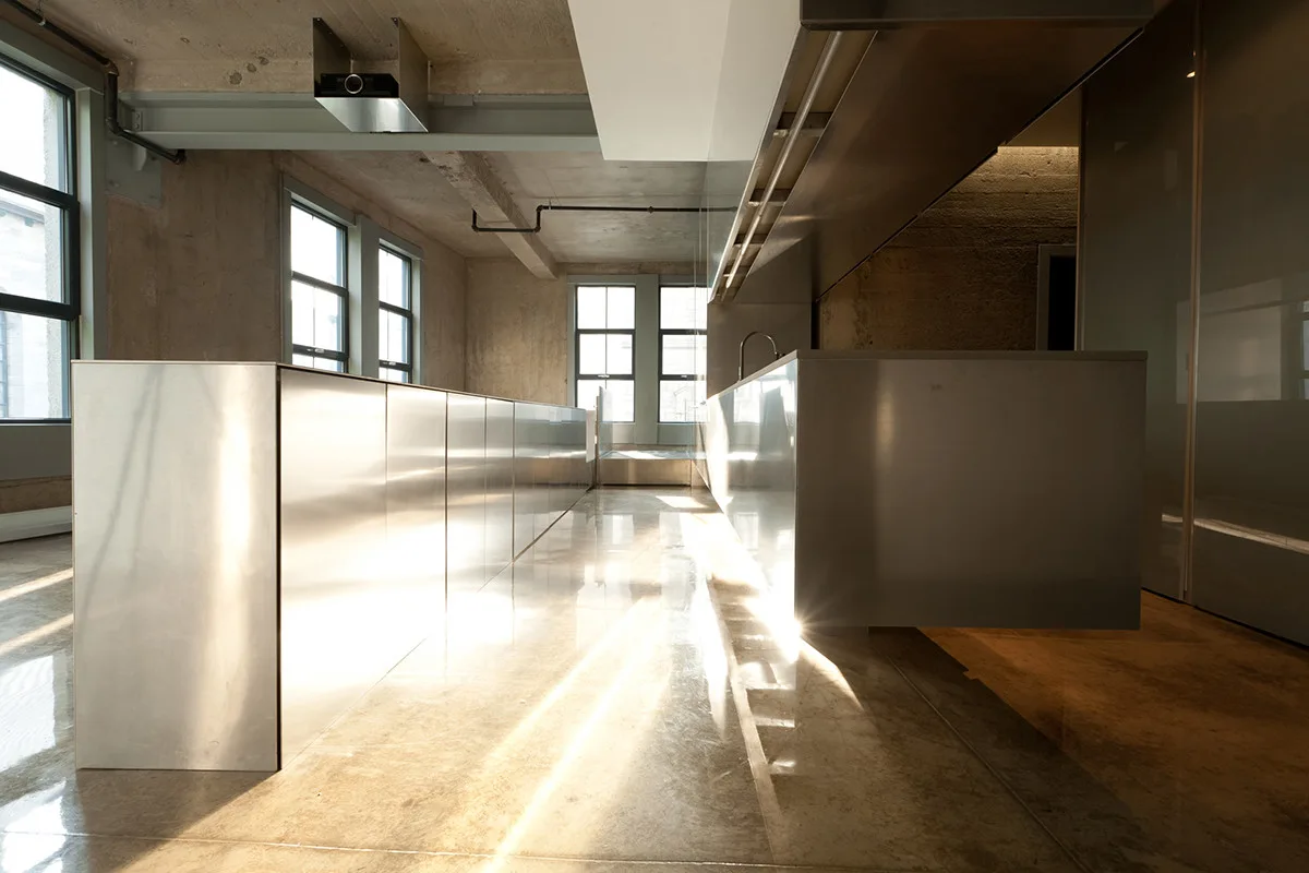 An industrial loft kitchen featuring a long stainless steel island, metal cabinets, polished concrete floors, and raw concrete walls.
