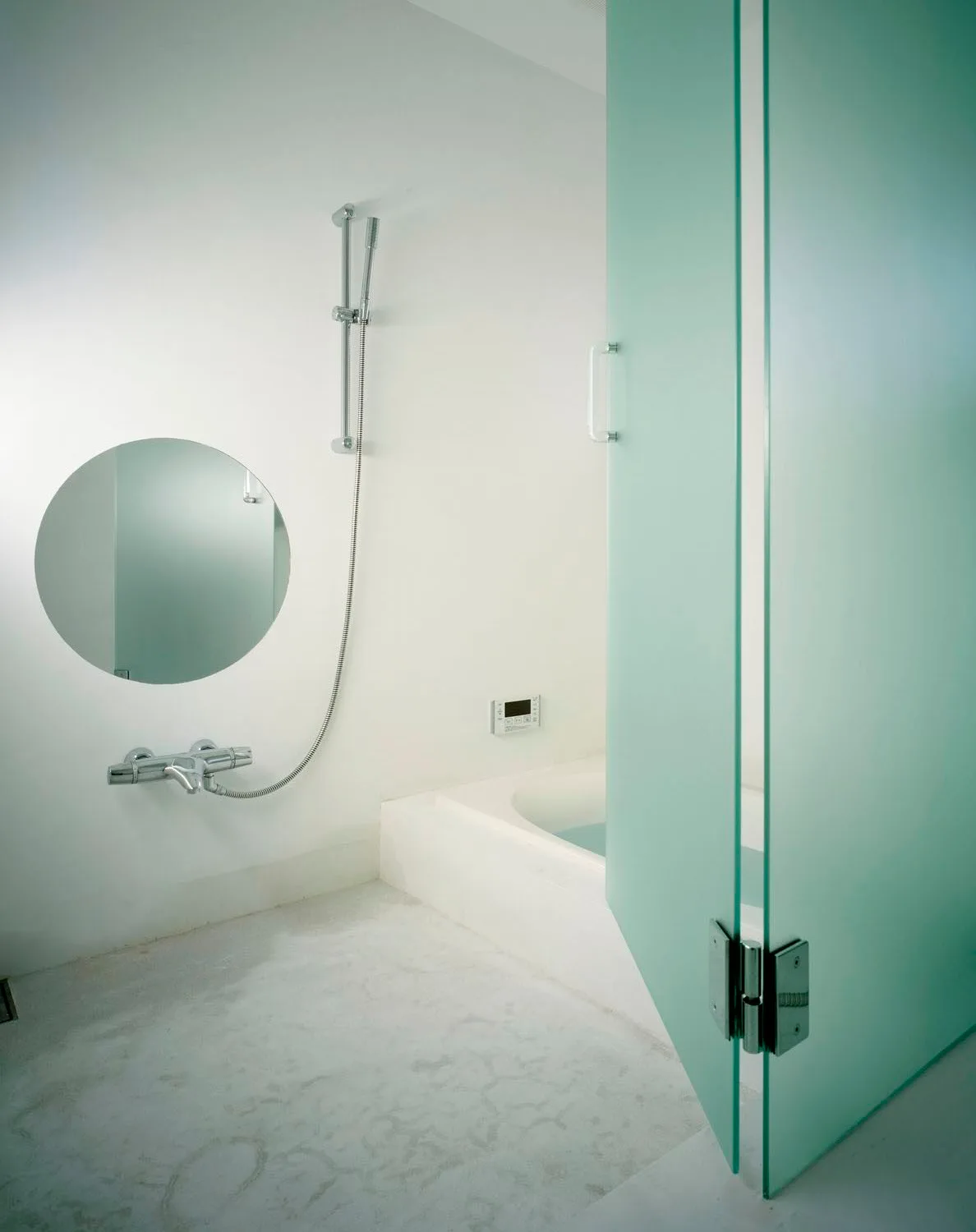 A minimalist white bathroom features a built-in tub, a circular wall mirror, chrome shower fixtures, and a large frosted mint green glass door.
