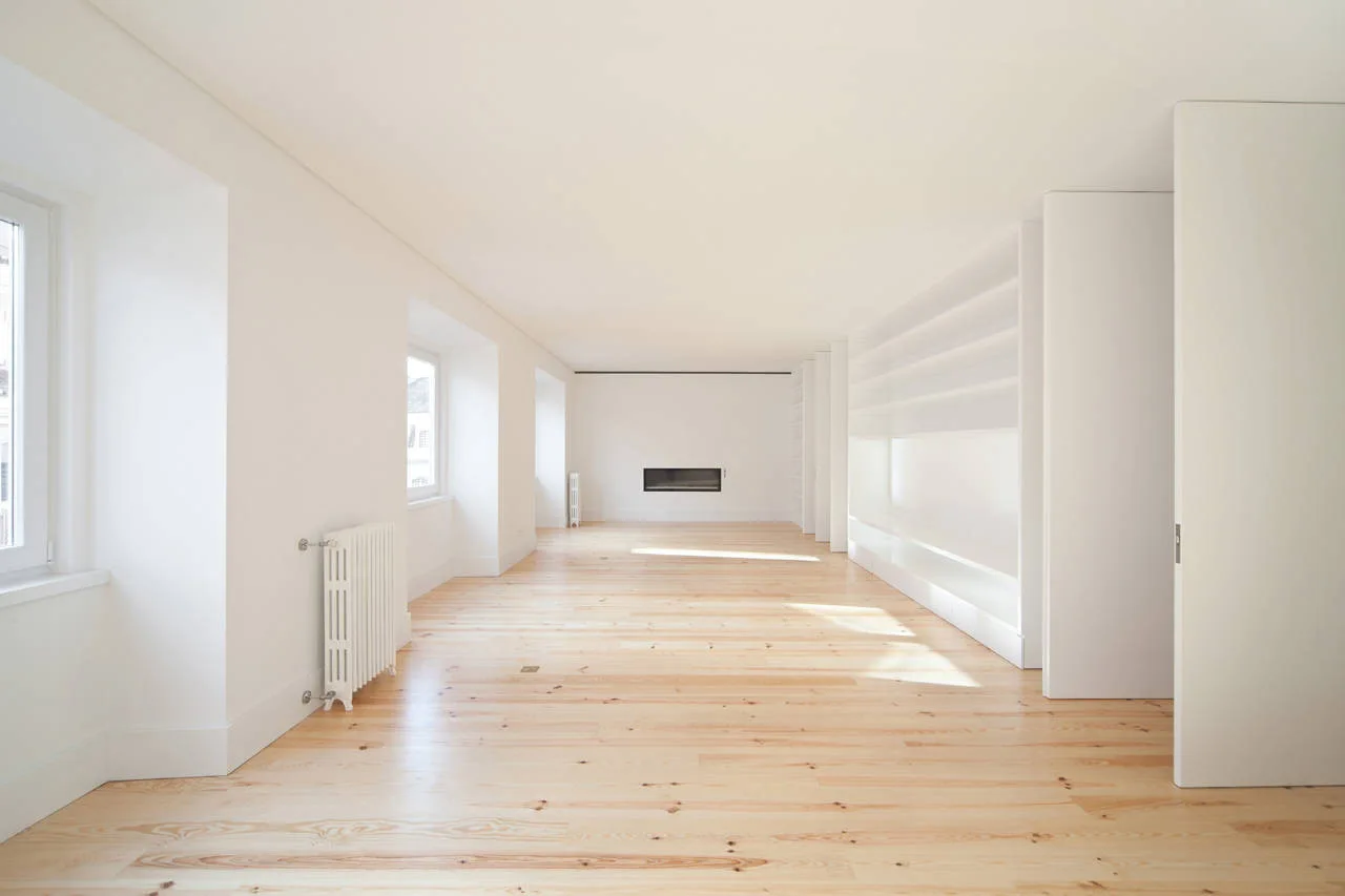 An empty minimalist living room featuring light wood plank flooring, white walls, deep recessed windows, and built-in white shelving.