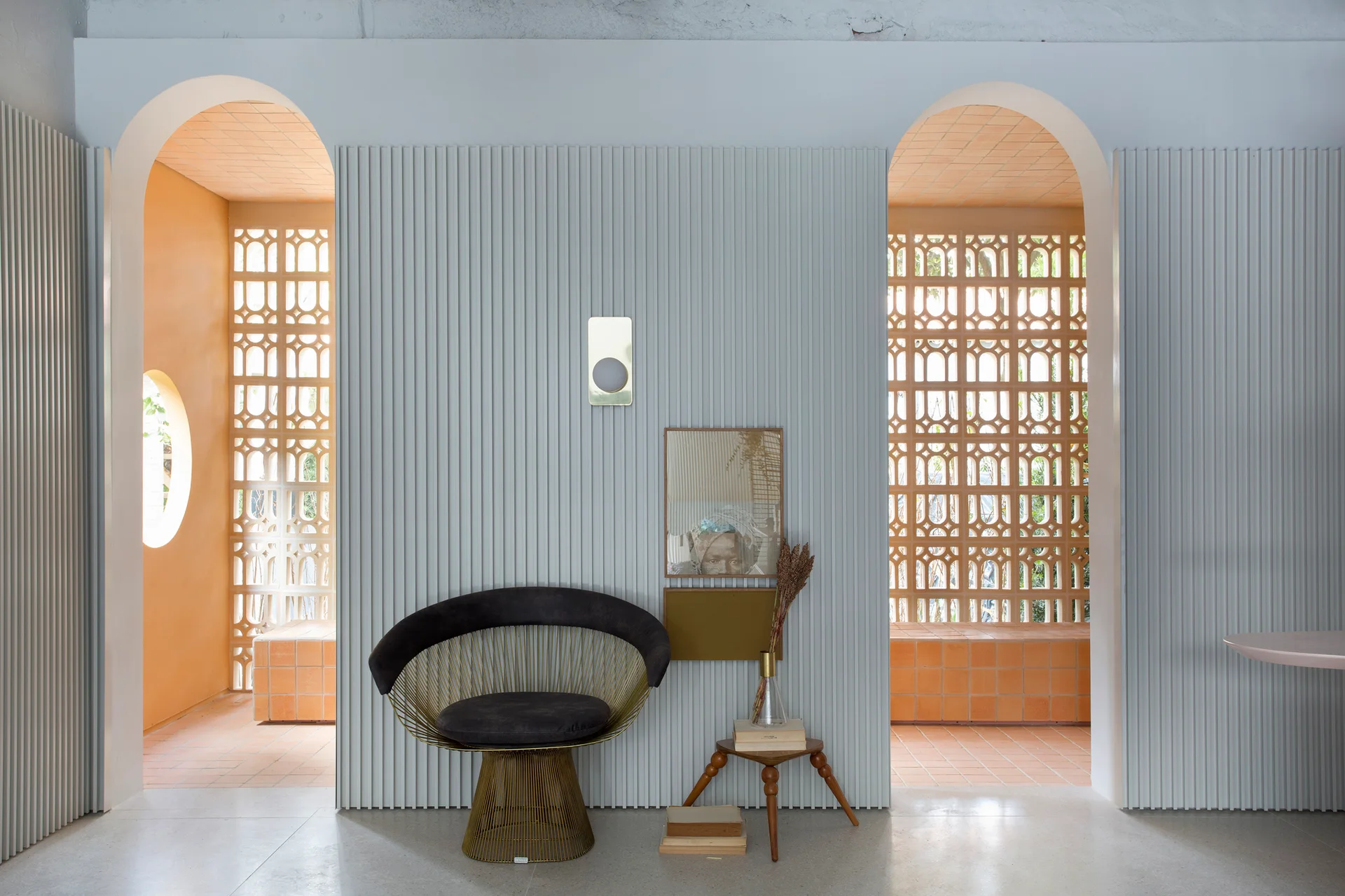 A modern black wire accent chair sits against a pale blue ribbed wall flanked by two arched doorways featuring decorative terra-cotta breeze blocks.
