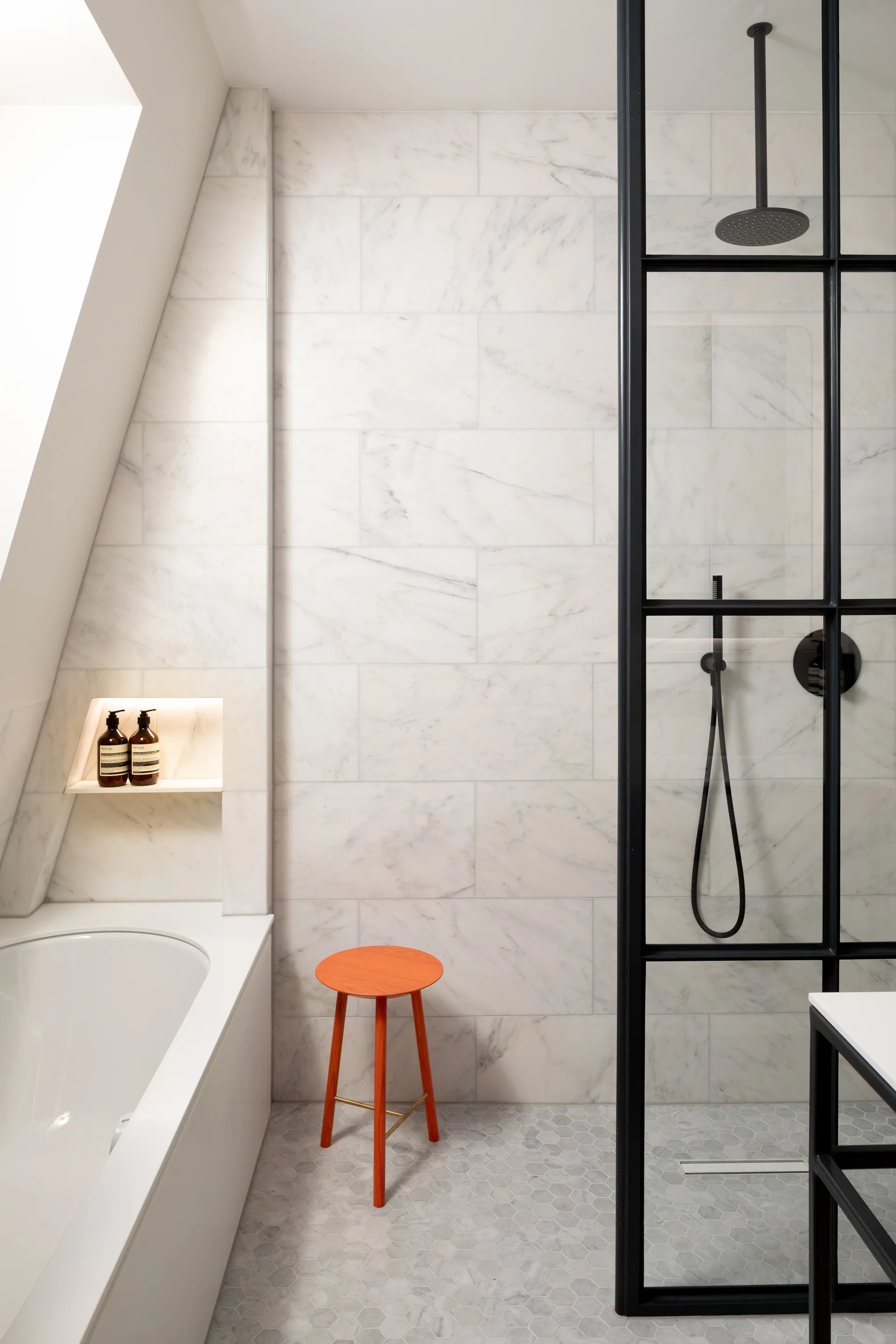A contemporary bathroom features marble wall tiles, a black-framed glass shower, a white bathtub, and a bright orange stool on hexagon floor tiles.