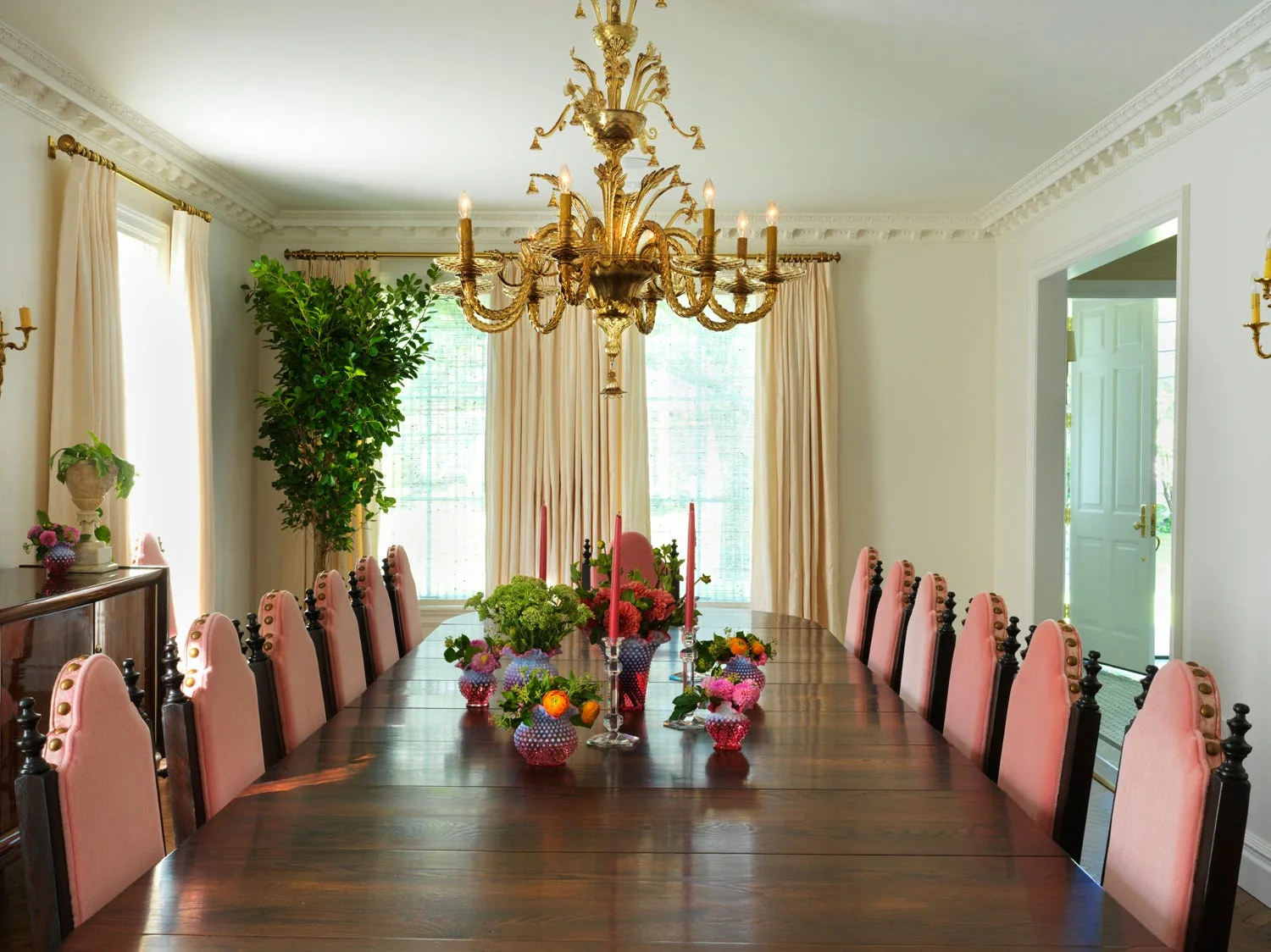 A formal dining room features a long dark wood table surrounded by pink upholstered chairs under an ornate gold chandelier.