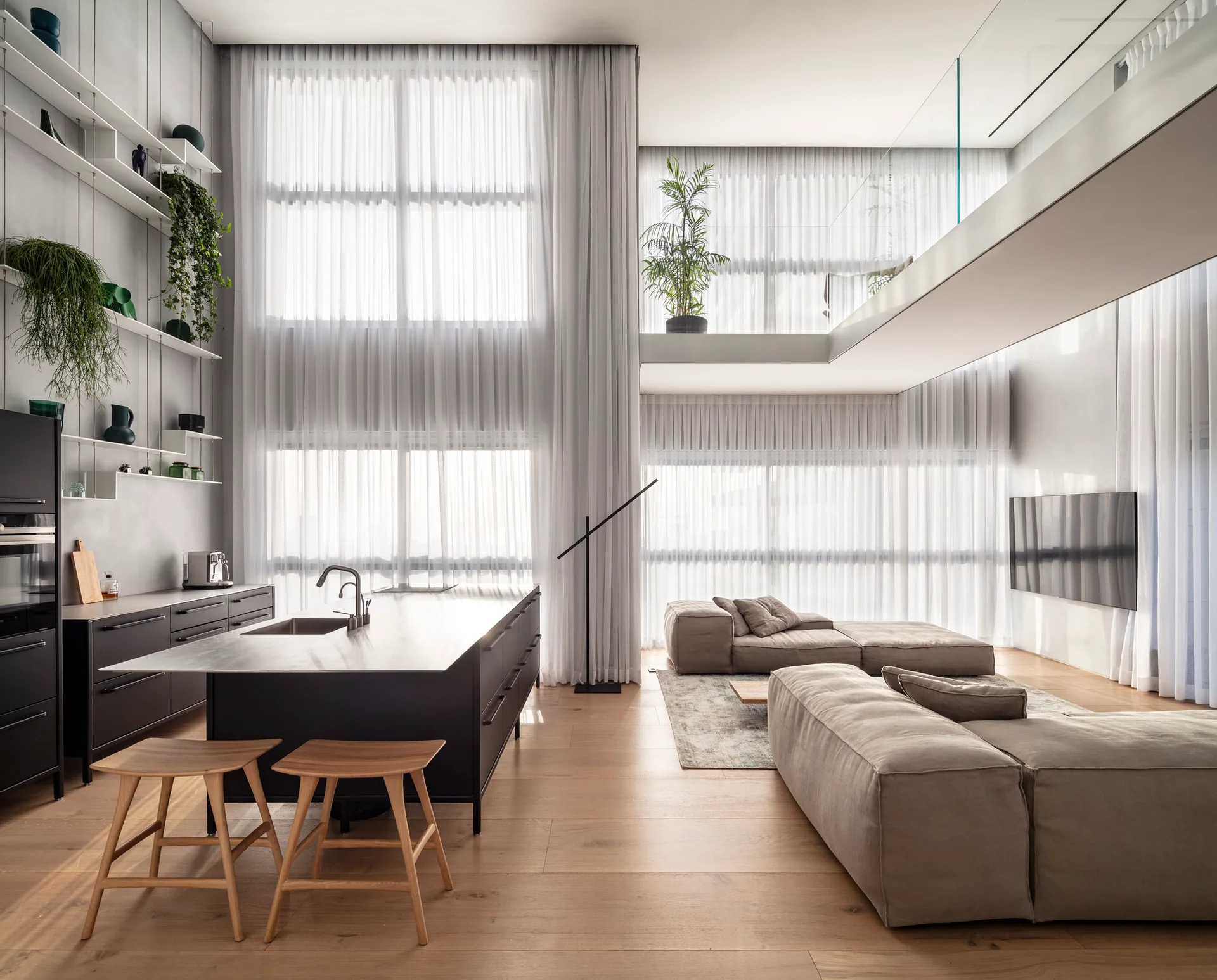 An open-plan living and kitchen space features a black island with wood stools, a modular sofa, and double-height sheer curtains.