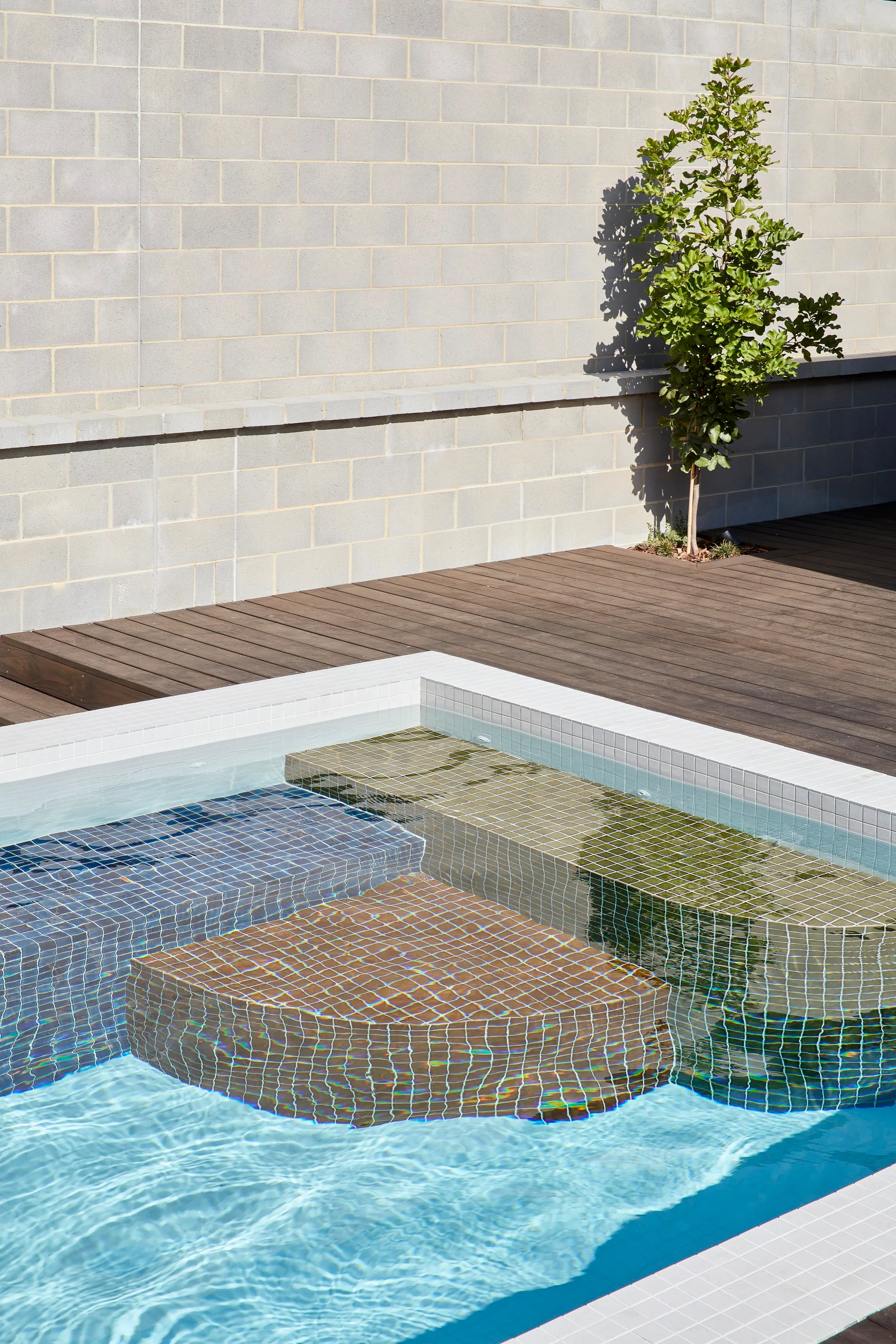 An outdoor swimming pool features blue, green, and brown mosaic tile steps next to a wooden deck and a grey masonry block wall.