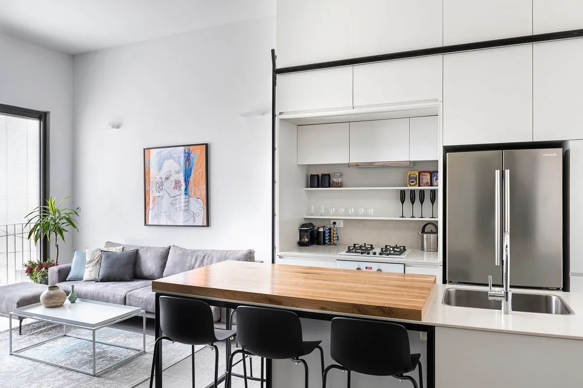 A modern open-plan kitchen and living room featuring white minimalist cabinetry, a wood-topped island with black stools, and a grey sofa.