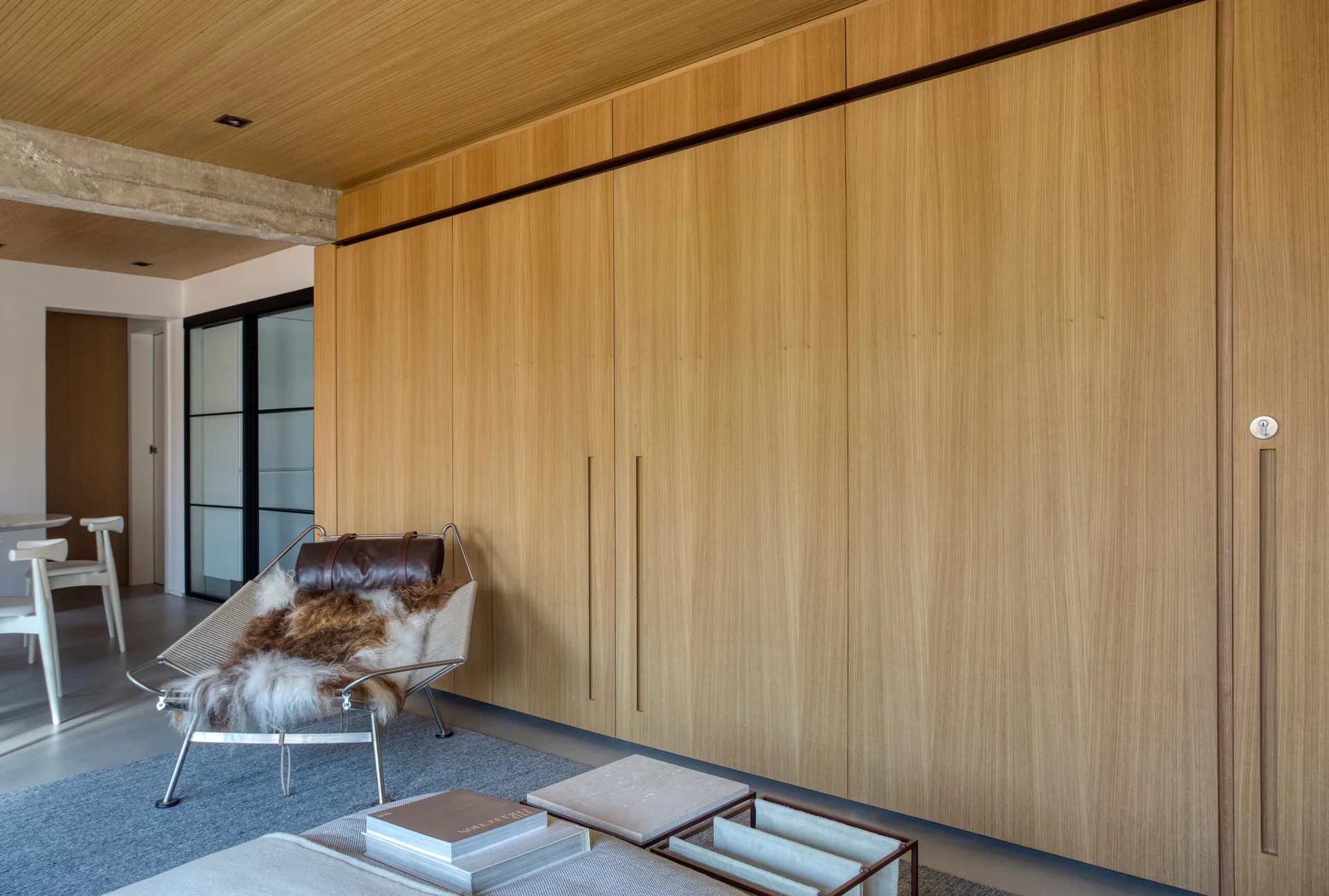 A modern living space features a massive floor-to-ceiling wood storage cabinet wall, a matching wood ceiling, and a metal lounge chair with a fur throw.