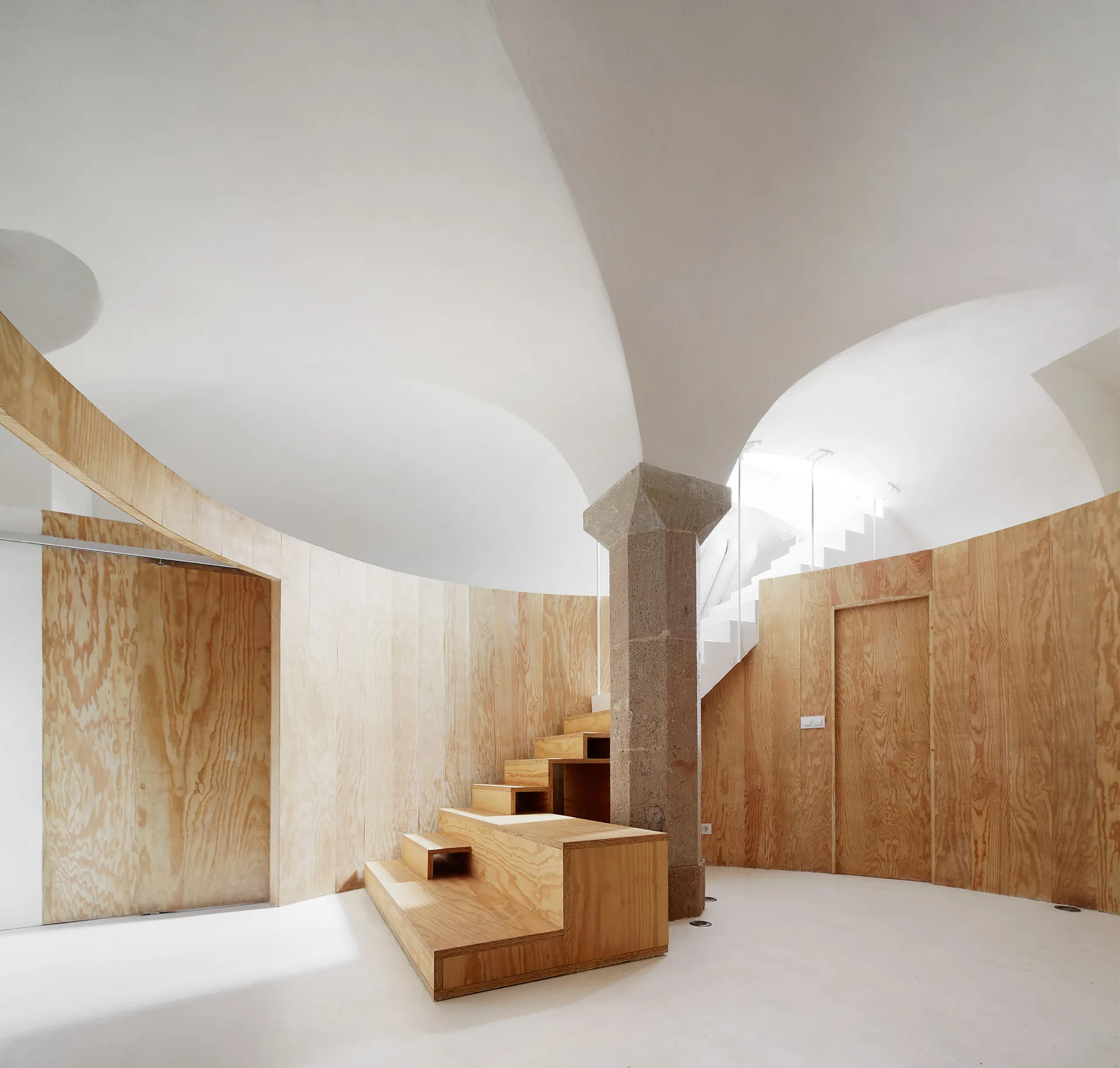 A minimalist interior space with a central wooden staircase, curved plywood wall panels, a gray stone pillar, and white vaulted ceilings.