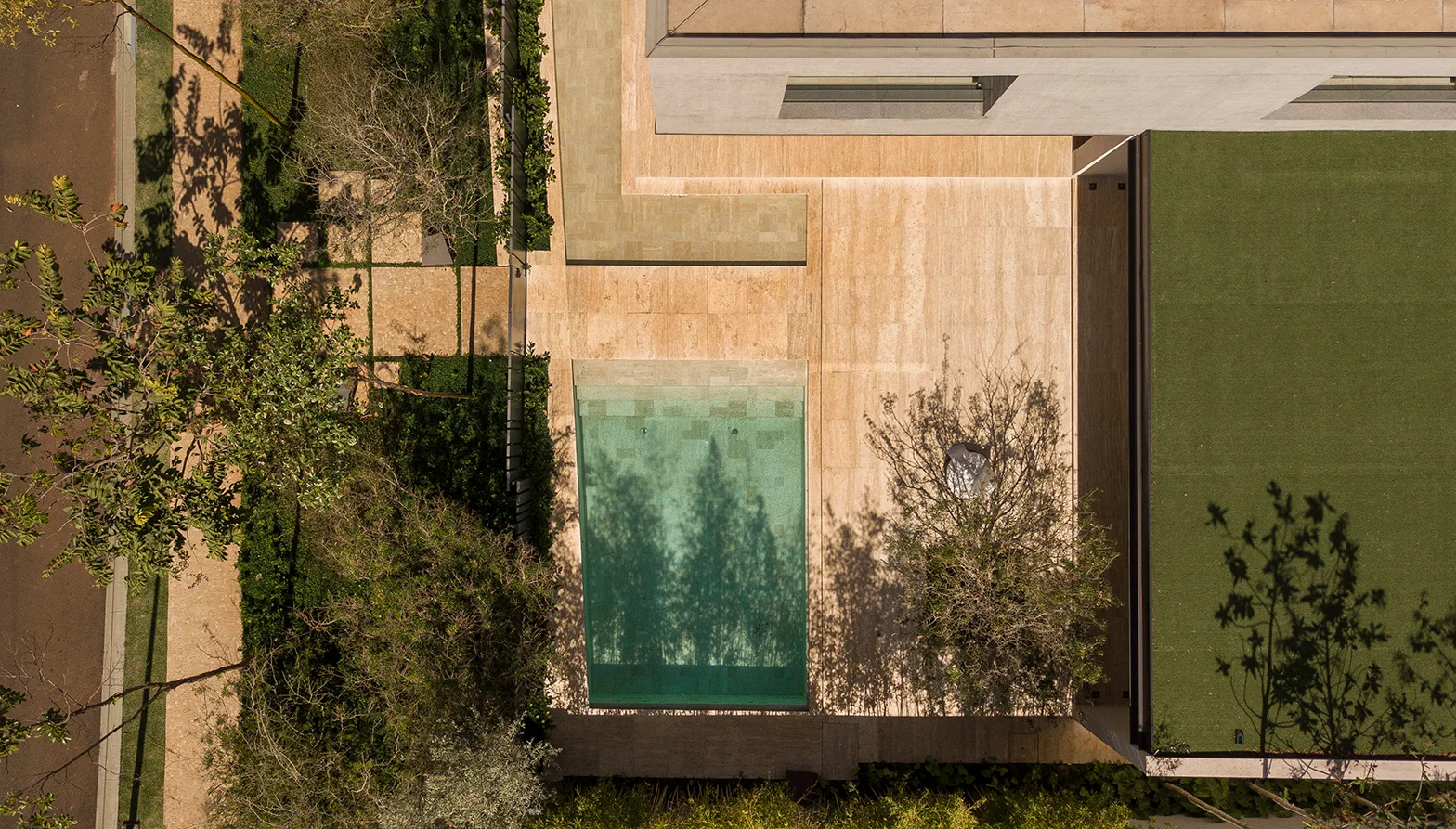 An aerial view of a modern backyard featuring a rectangular swimming pool, beige stone patio paving, green lawn, and trees casting shadows.