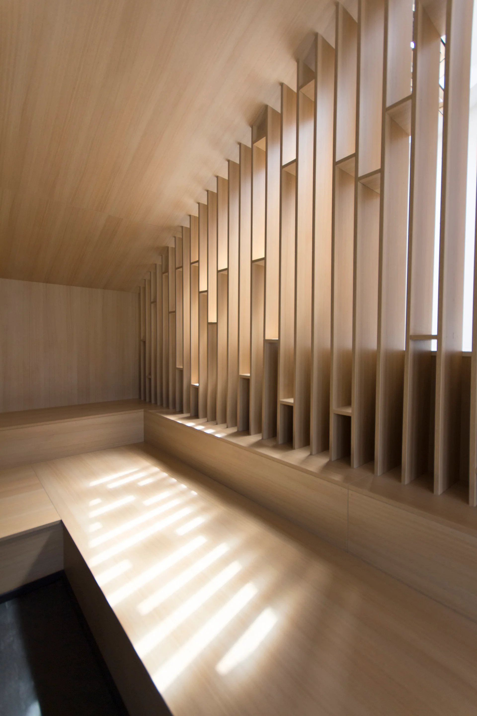A minimalist wood-paneled room features a sloped ceiling, an integrated bench, and a tall vertical slatted partition filtering natural sunlight.
