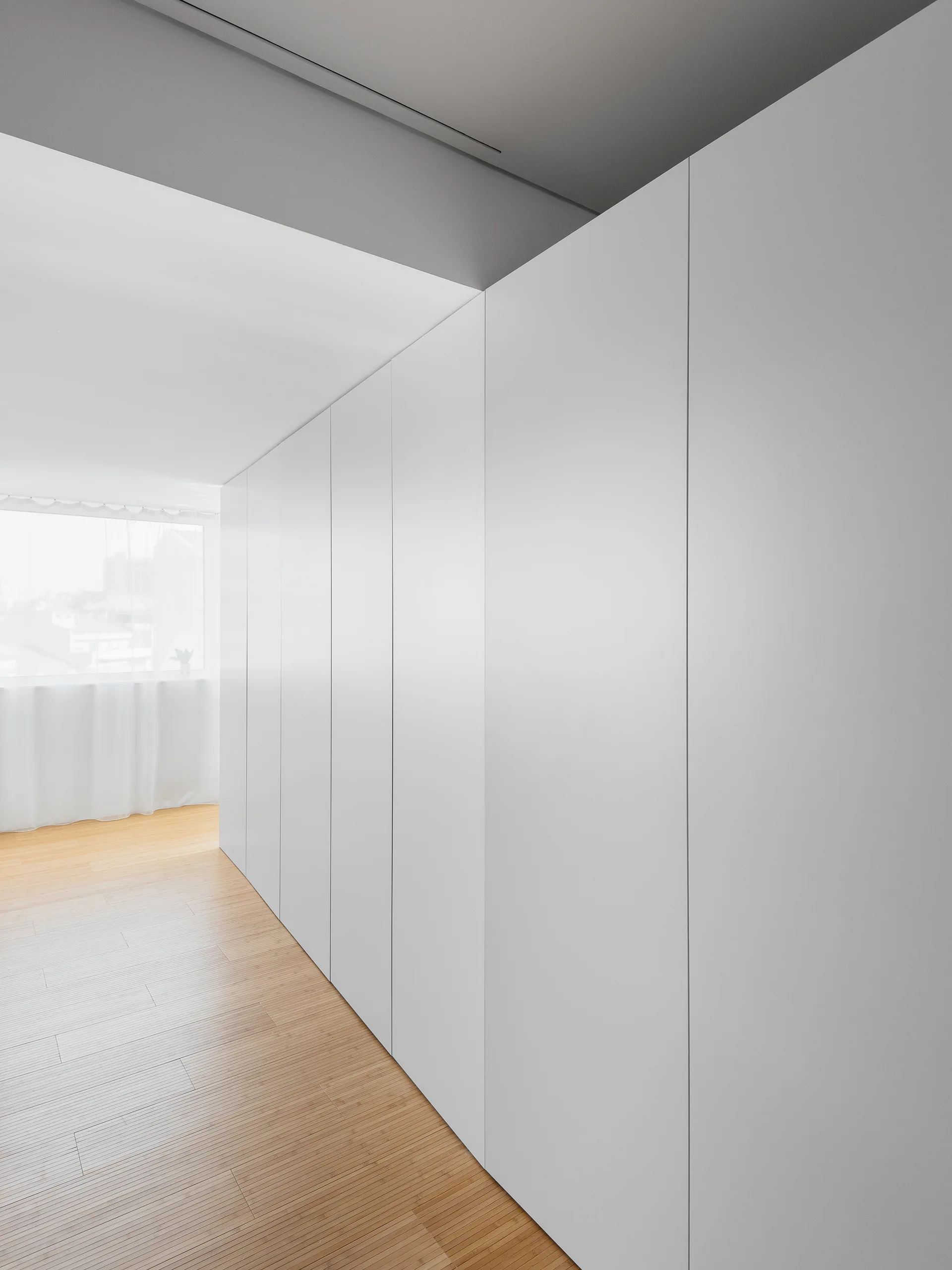A minimalist hallway features a floor-to-ceiling wall of matte white handleless storage cabinets beside a light hardwood floor leading to a window.