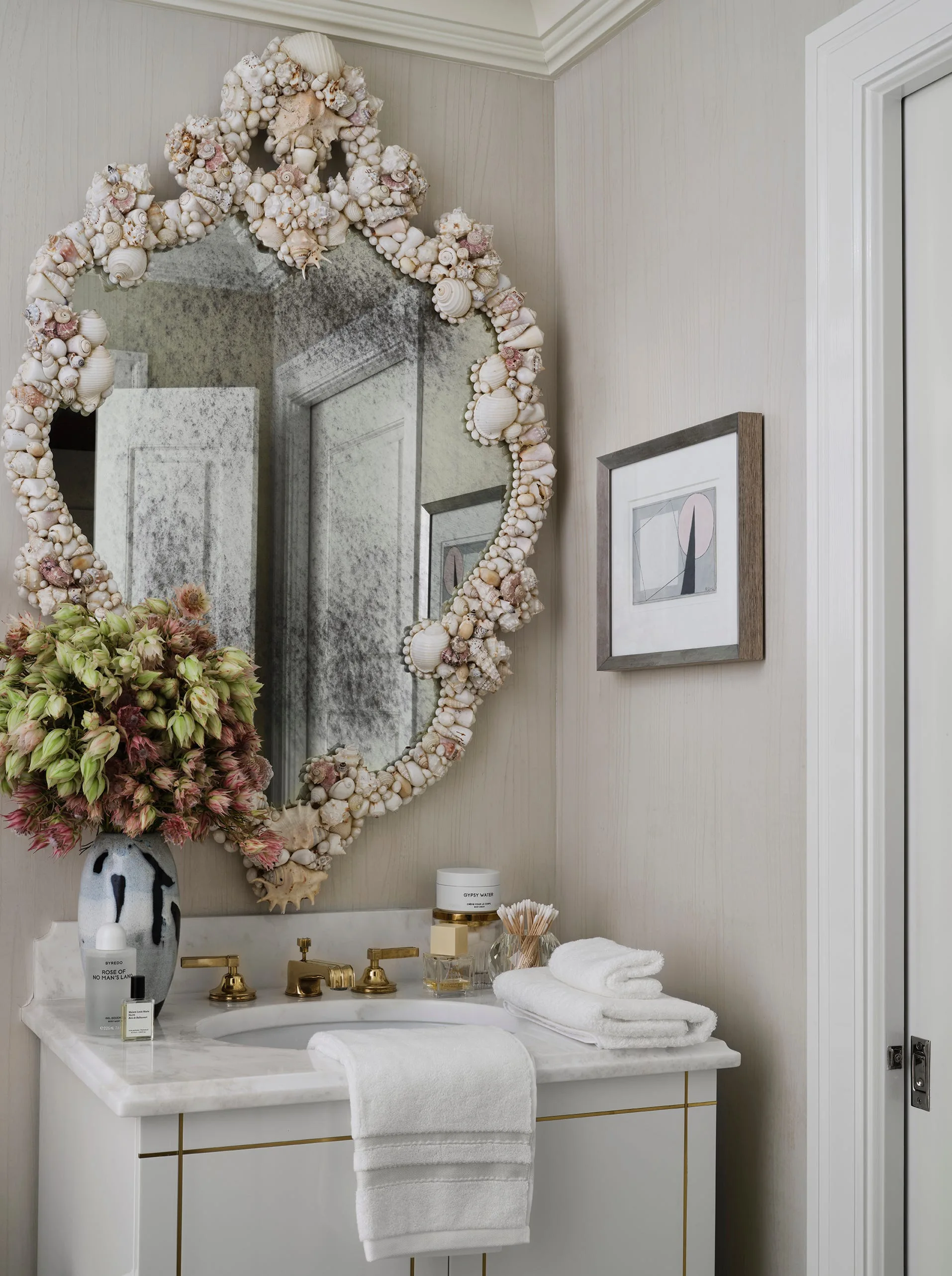 A bathroom vanity with a white marble countertop, gold faucet, and a large mirror framed in hundreds of small white seashells.