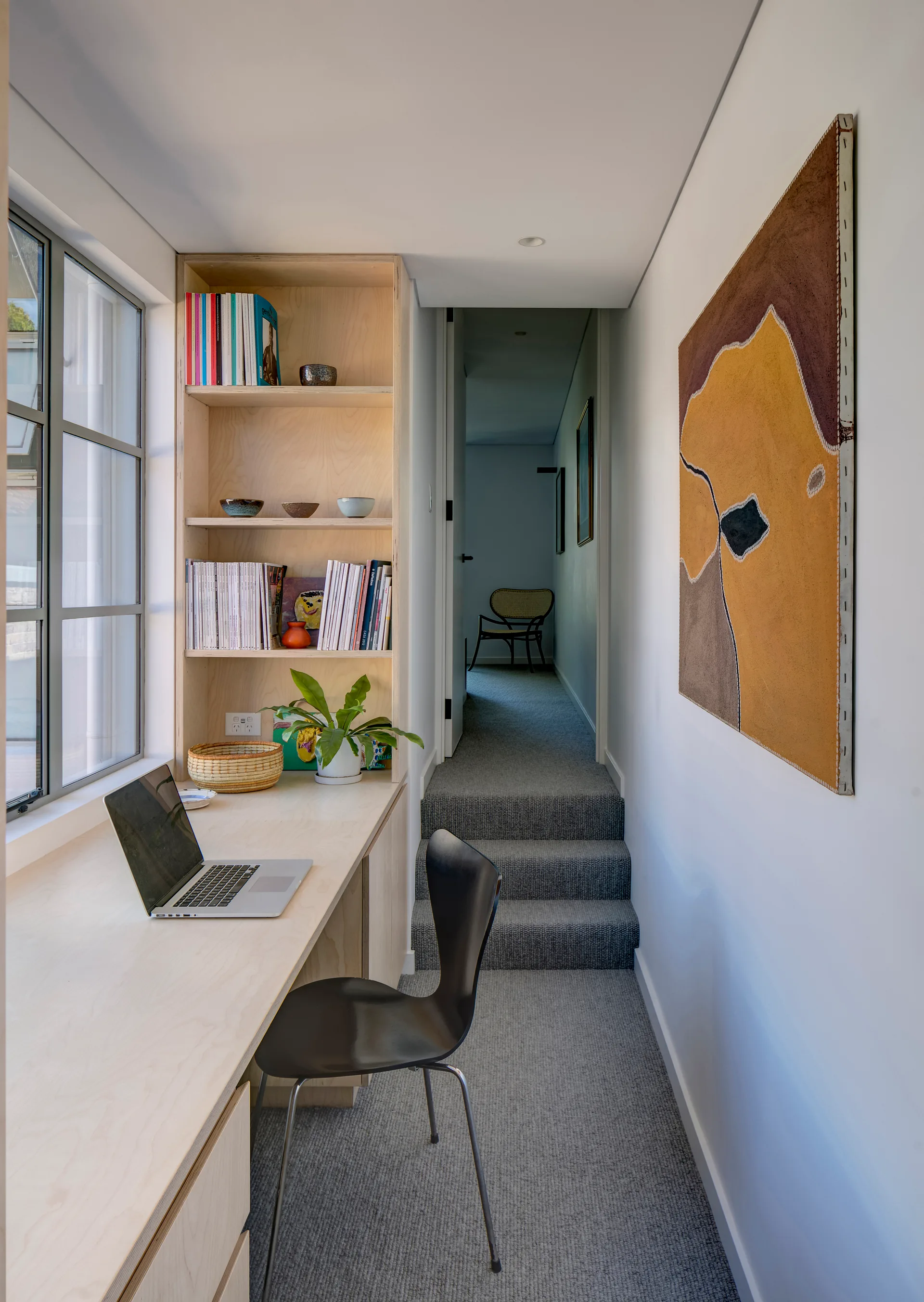 A compact workspace features a built-in plywood desk and shelving unit next to a grid window and carpeted stairs leading to a hallway.