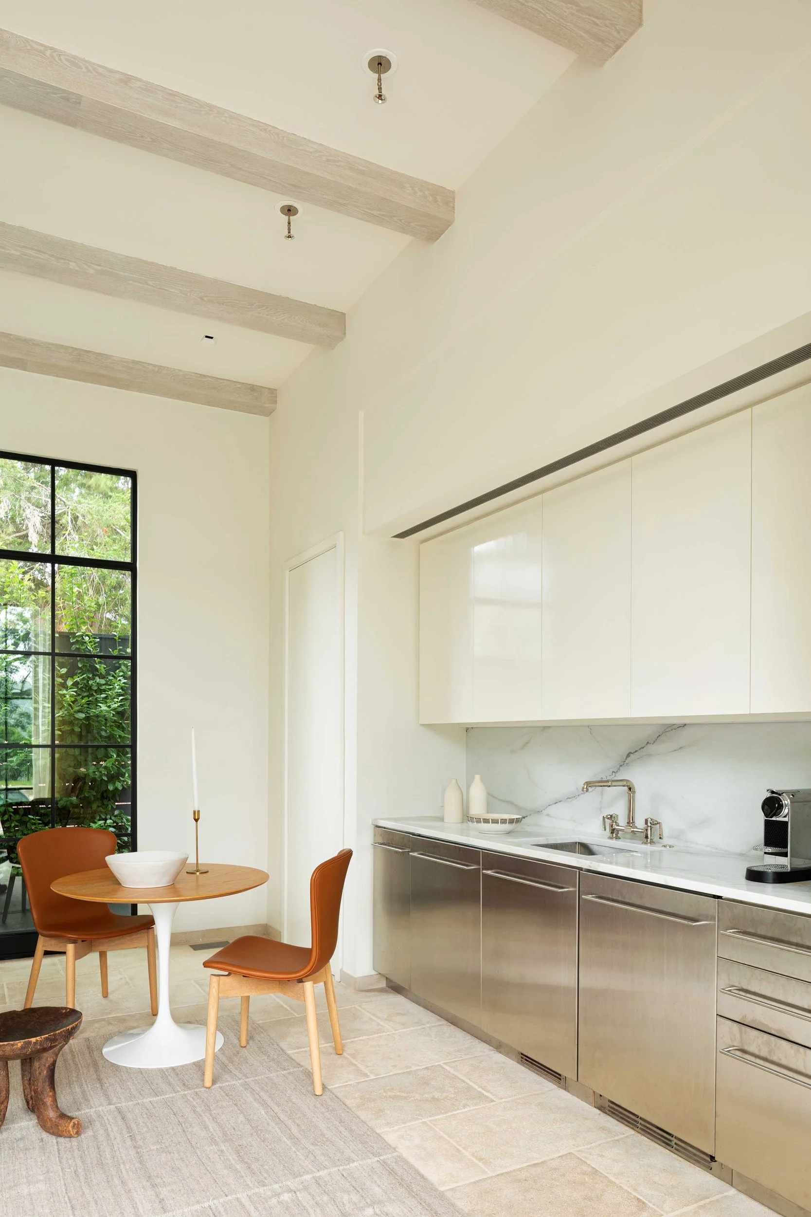 A modern kitchen with stainless steel base cabinets, white marble backsplash, exposed wood ceiling beams, and a small dining table with leather chairs.