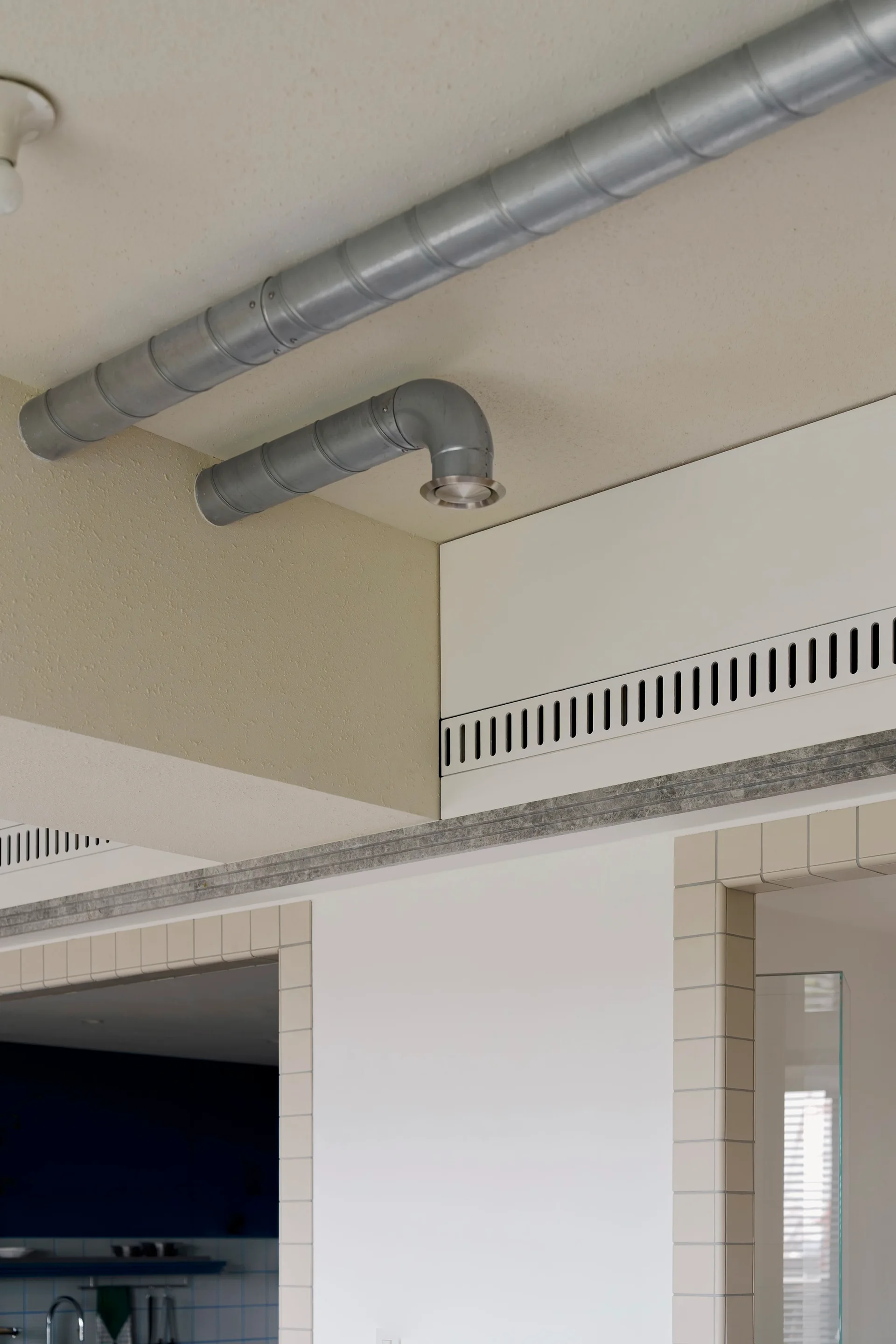 Exposed metal HVAC ducts run along a beige ceiling above white walls with tiled doorway casings and a slotted ventilation panel.