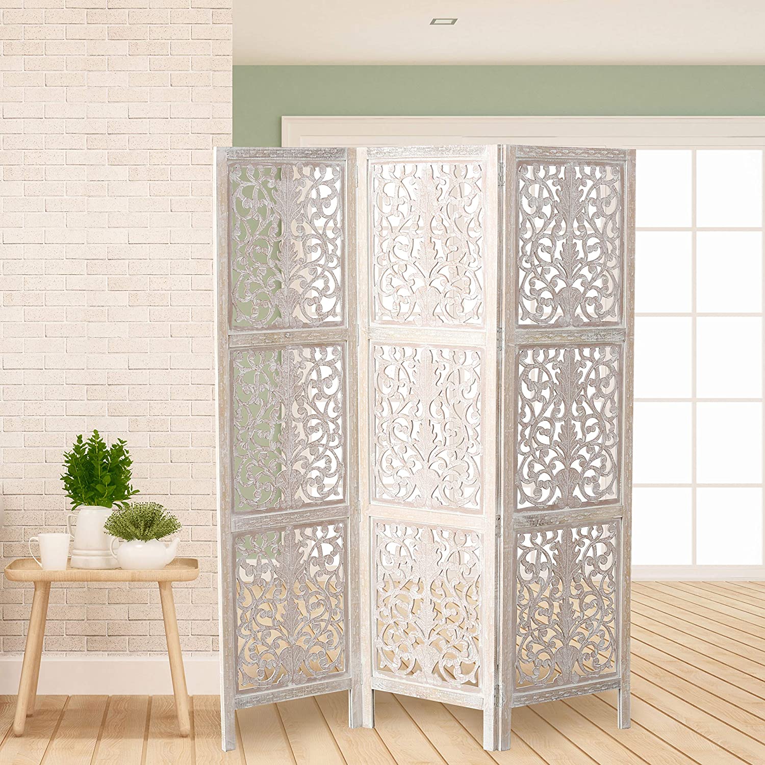 A three-panel white-washed wood folding screen with intricate floral carvings stands on a light hardwood floor next to a white brick wall.