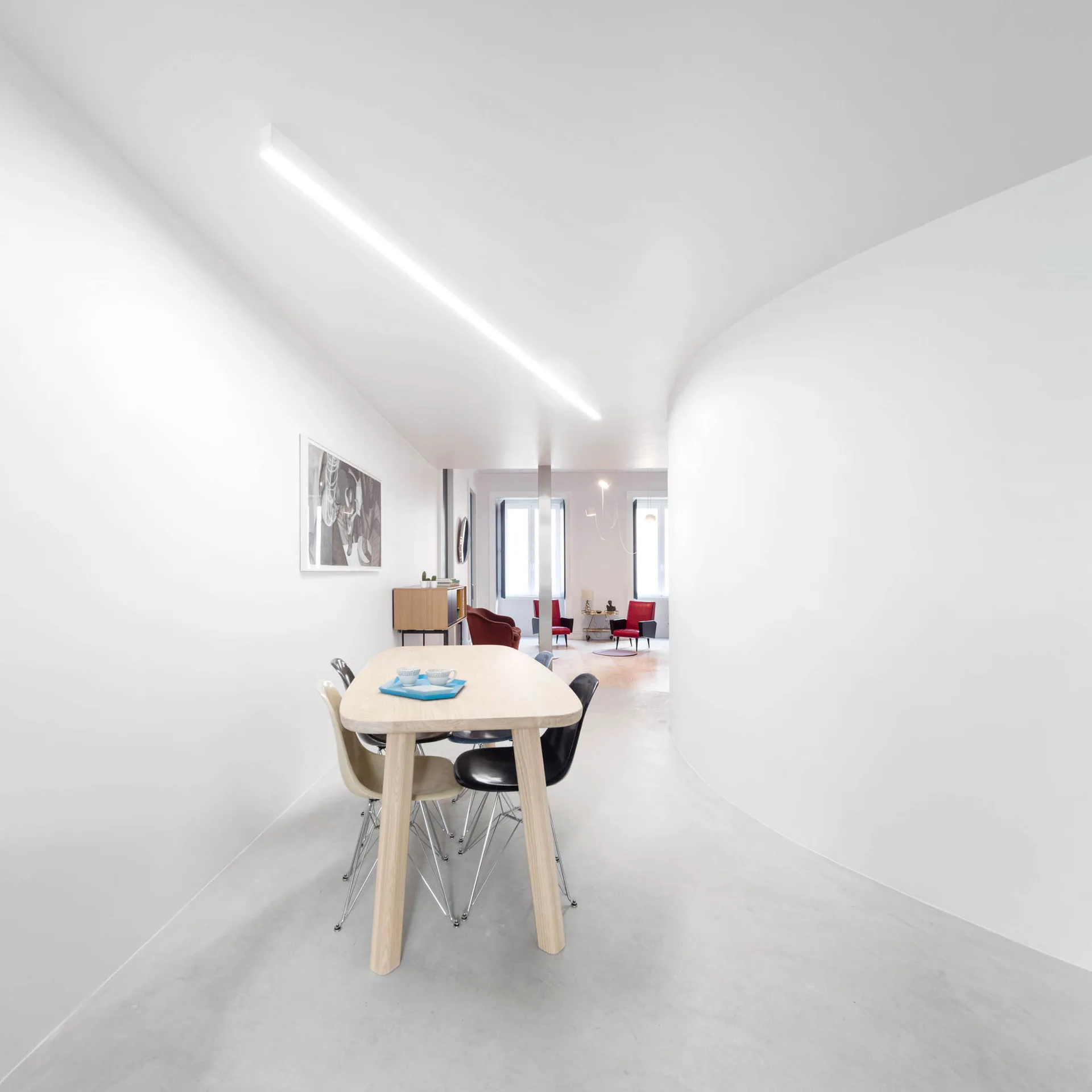A minimalist dining area features a light wood table with mid-century chairs set against a curved white wall and a long linear ceiling light.
