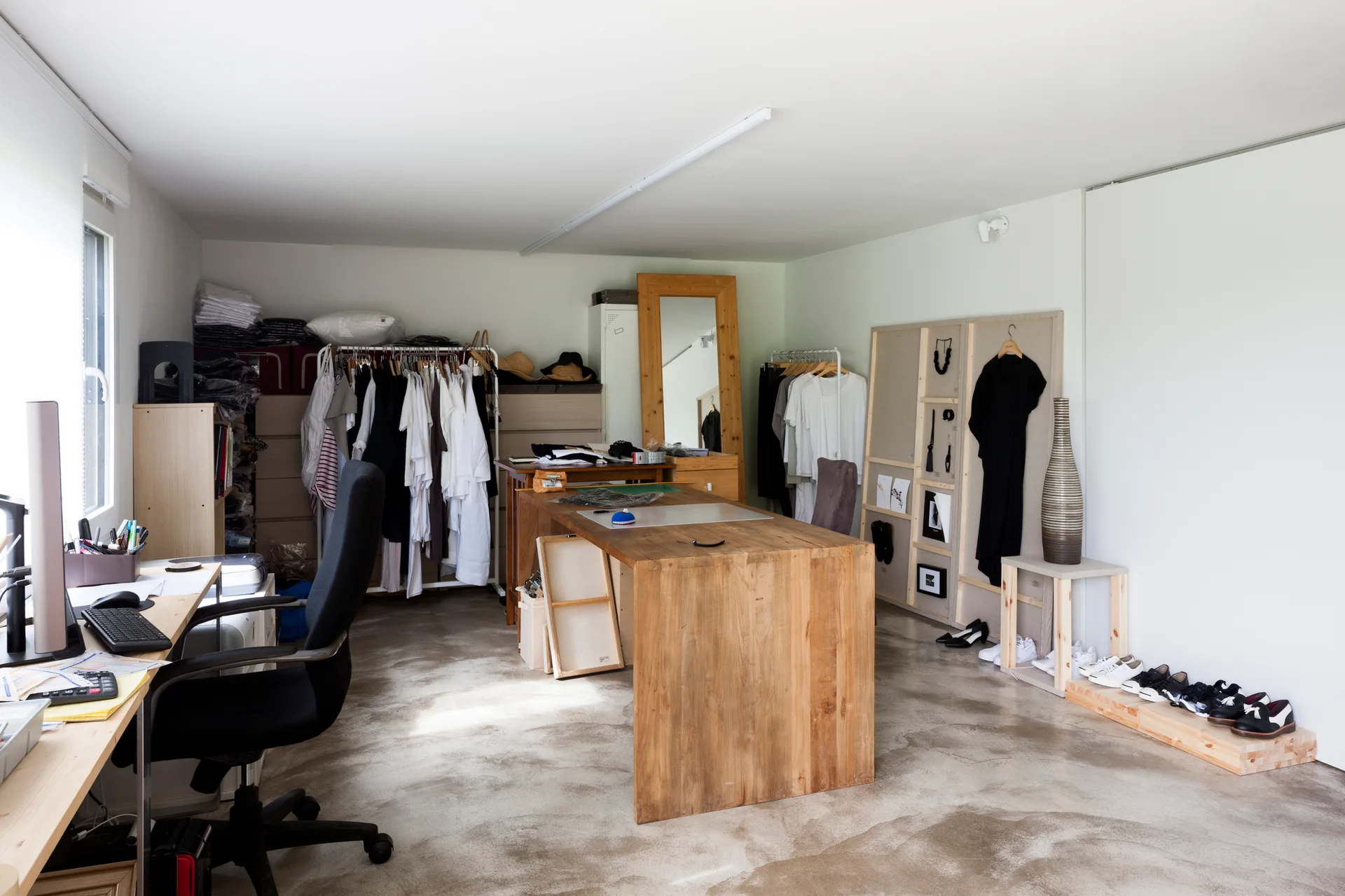 A minimalist fashion studio featuring raw concrete floors, large wooden work tables, clothing racks, and a full-length wooden floor mirror.
