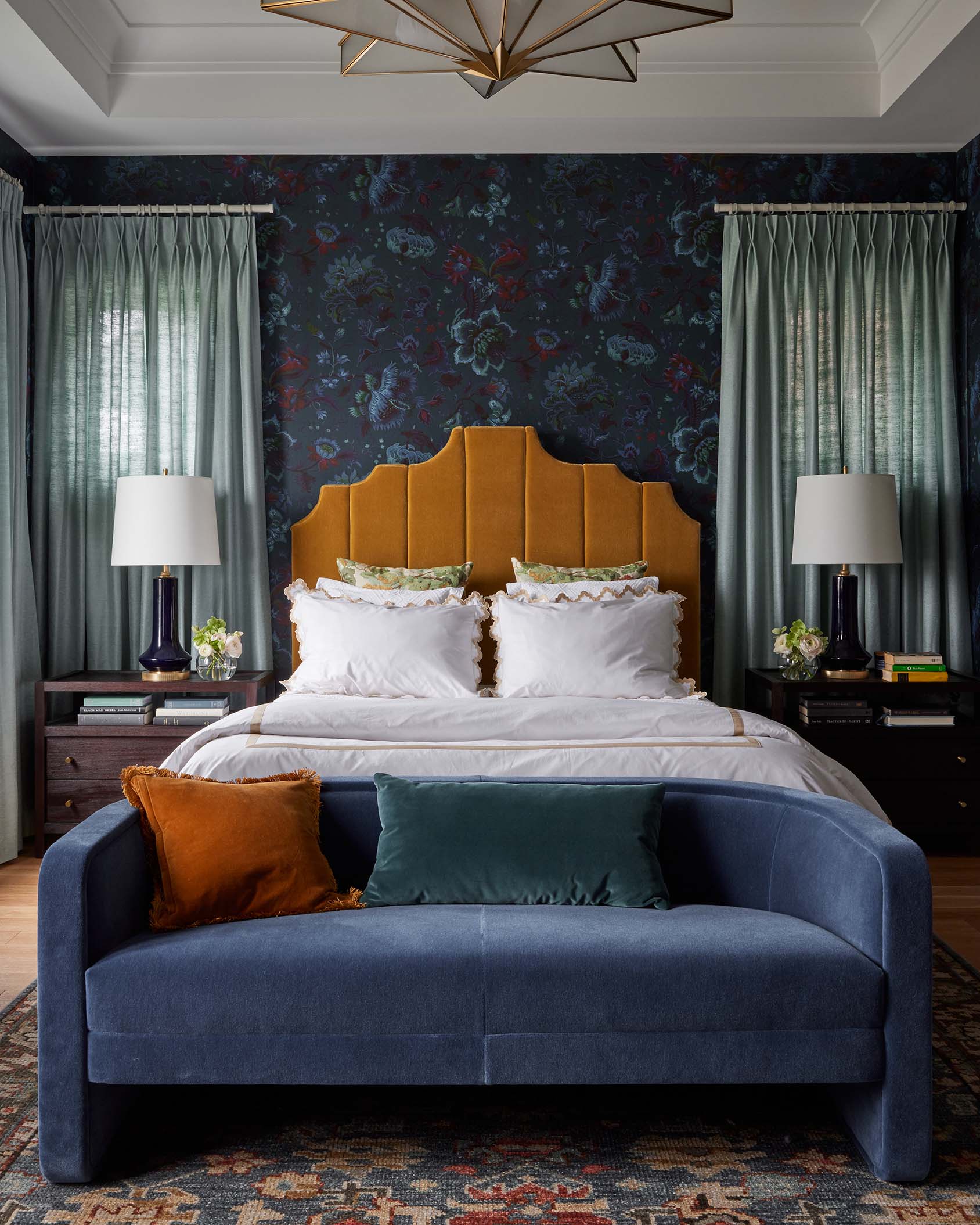 A symmetrically arranged bedroom features a mustard yellow headboard against dark floral wallpaper, with a curved blue sofa placed at the foot of the bed.
