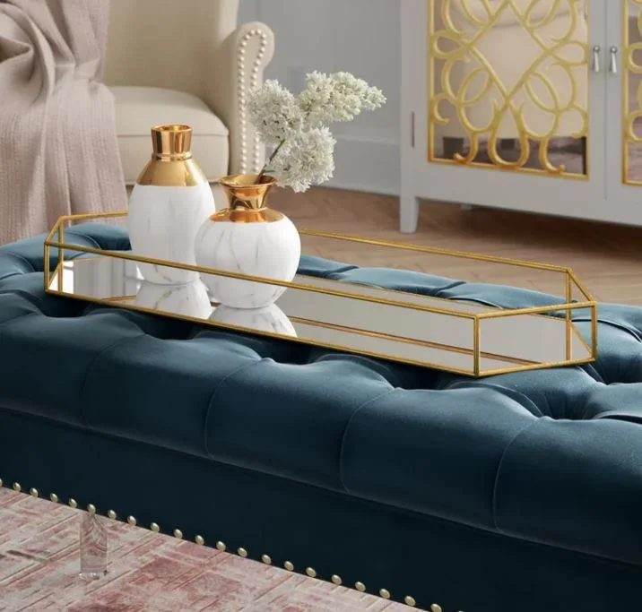 A gold mirrored tray with two marble-patterned vases sits on a tufted blue velvet ottoman.