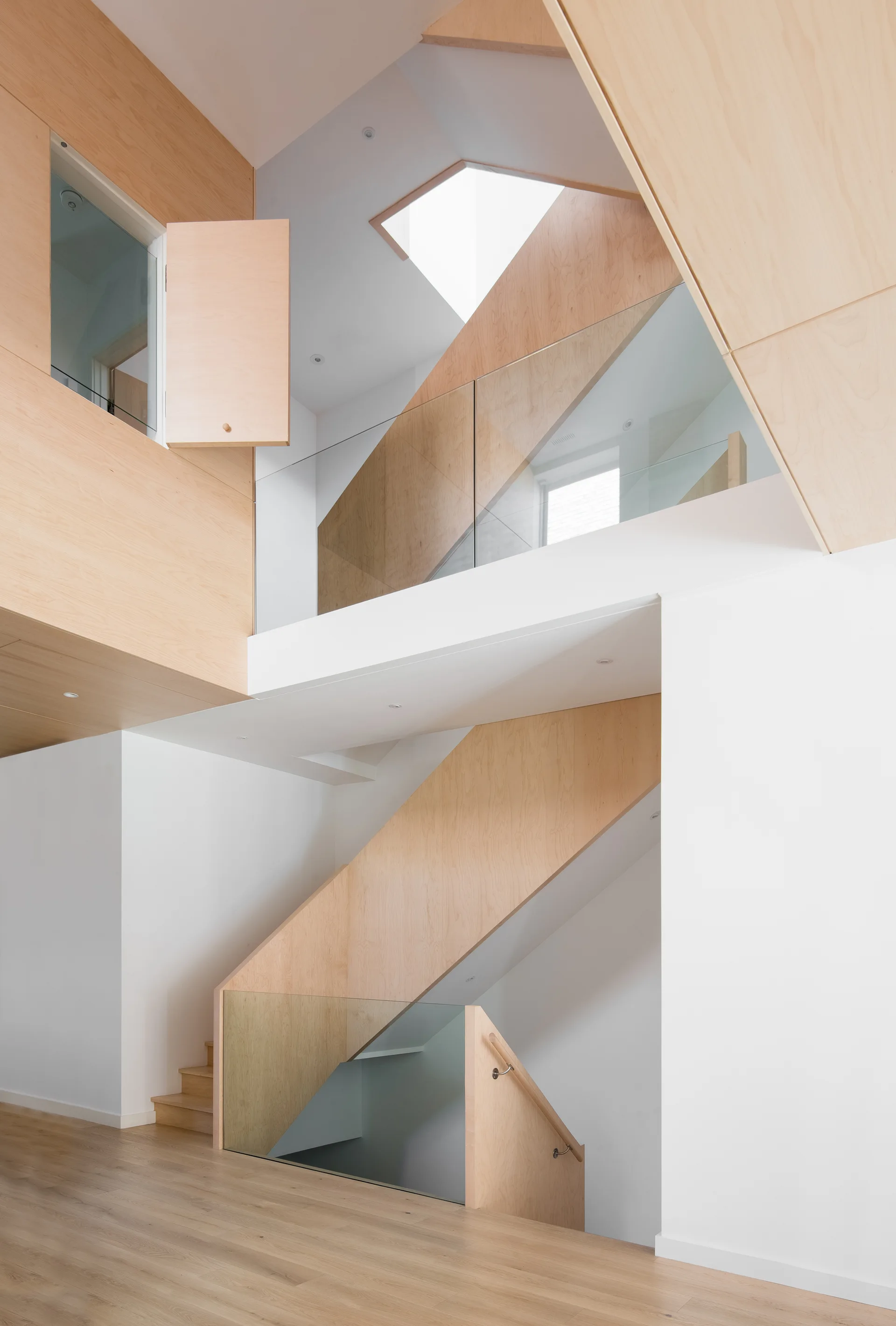 A modern multi-level staircase with light wood plywood wall panels, clear glass railings, and a geometric ceiling skylight.