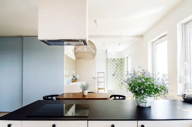 A minimalist kitchen features a black island countertop, white range hood, pale blue sliding door, and a glimpse of green checkered bathroom tile.