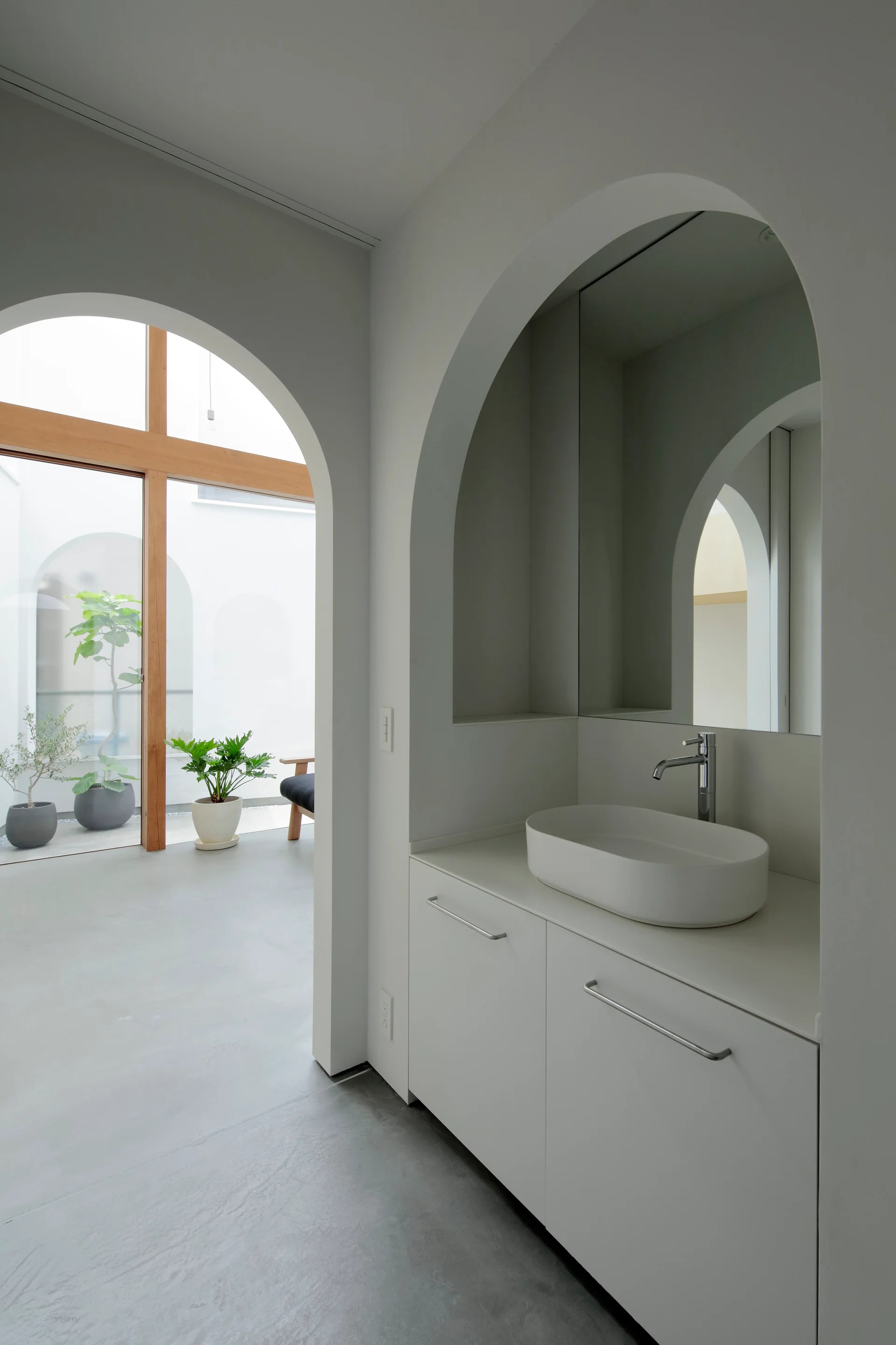 A minimalist white vanity with a vessel sink is nested inside an arched alcove next to a doorway looking out to a bright courtyard.