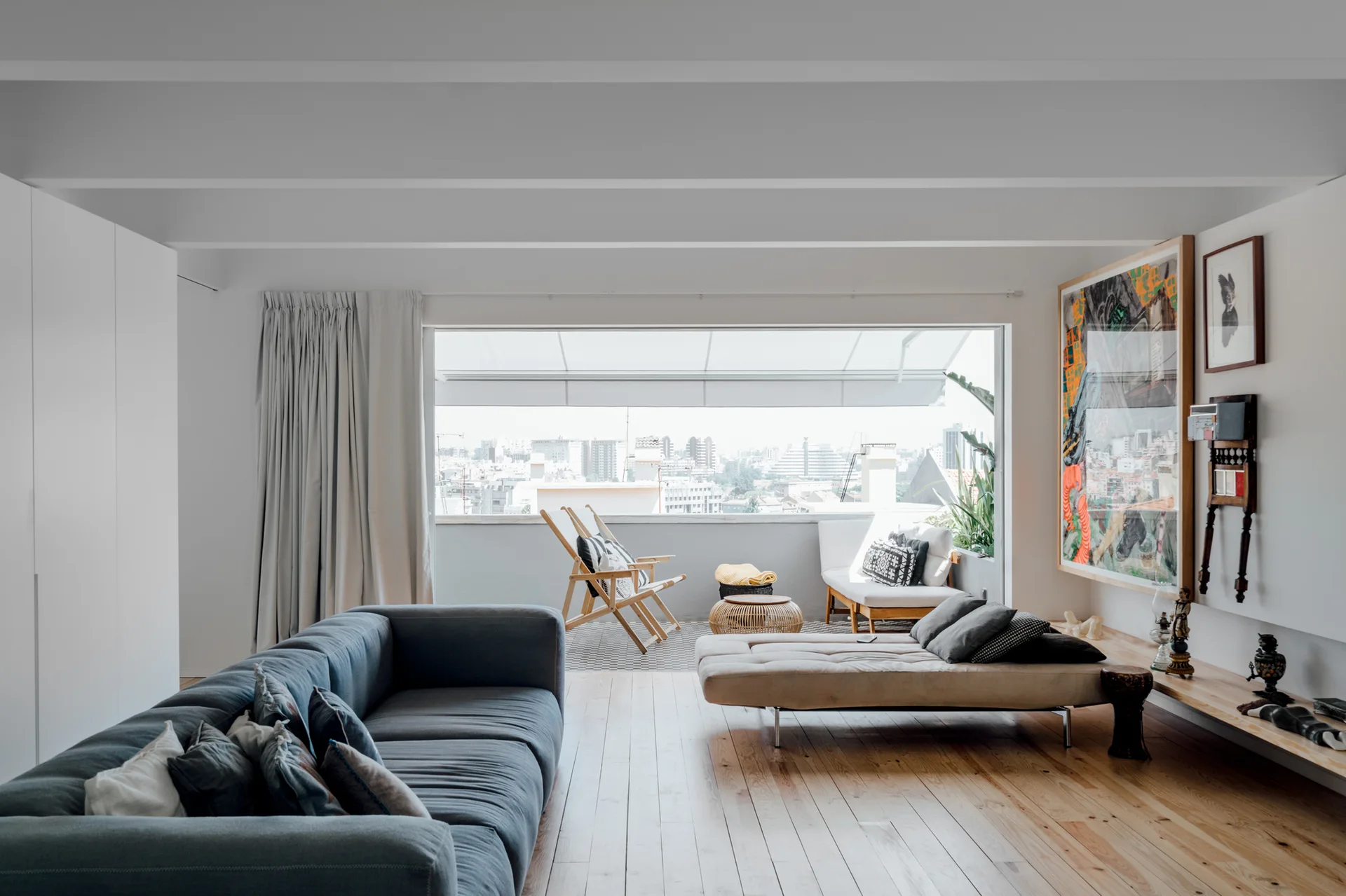 A bright living room with a grey sofa, a beige daybed, light wood floors, and a wide window opening to a balcony with city views.